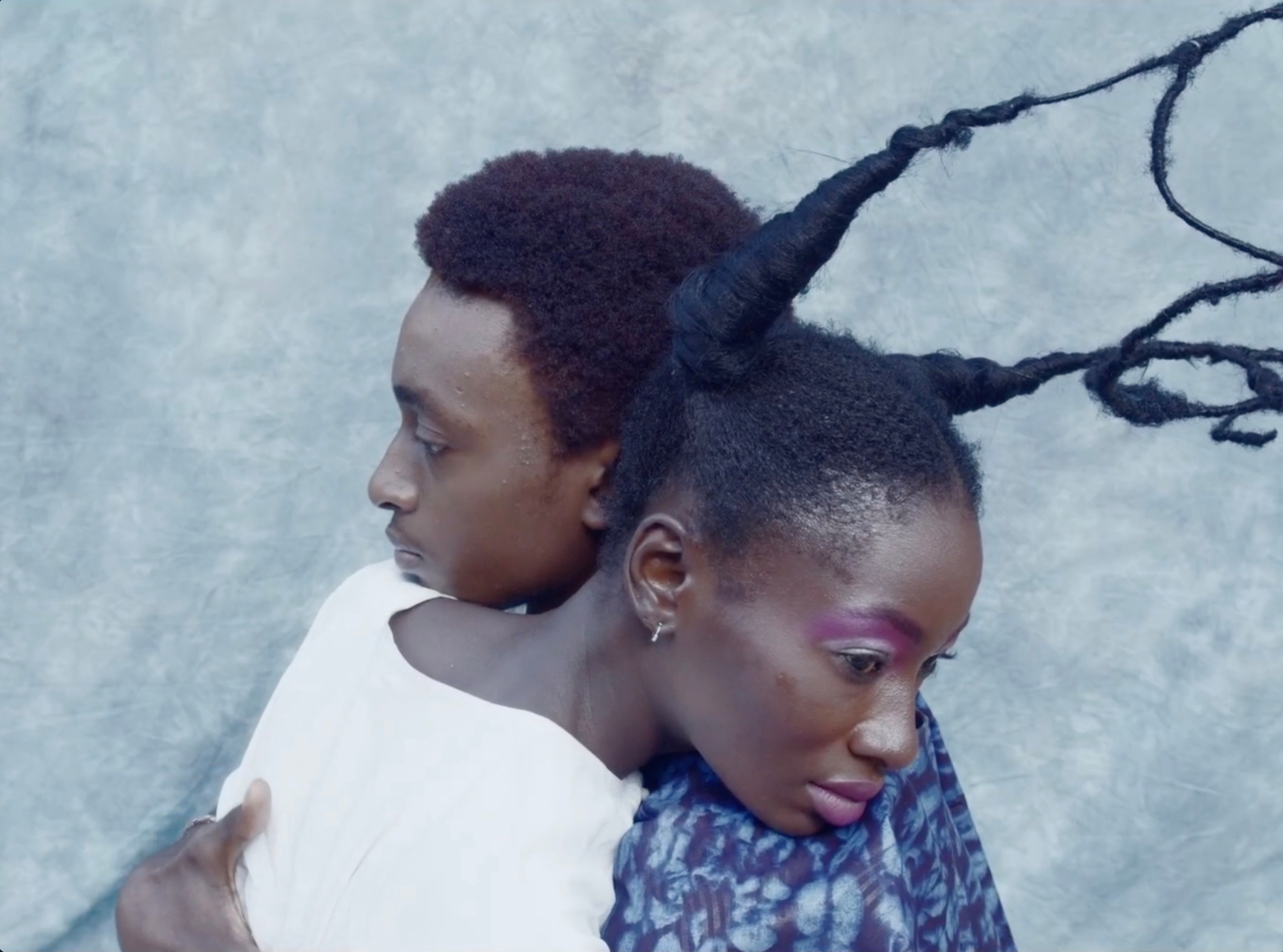 Two people are embracing against a soft, cloudy grey backdrop. The person on the left, wearing a white garment, faces sideways with a calm expression, their short, textured hair slightly damp with sweat. The person on the right leans into the hug, wearing a patterned blue and purple top, with vibrant pink eyeshadow and lipstick. Their hair is styled into two long, twisted, and sculptural spikes extending outward, giving a bold, artistic look. The mood appears emotional and intimate.