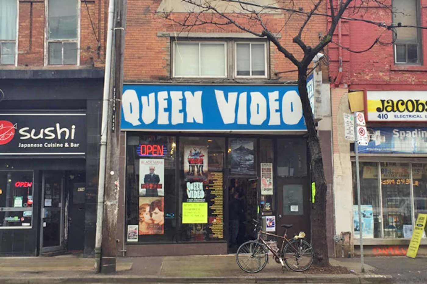 A streetscape featuring three storefronts. In the centre is “QUEEN VIDEO,” a video rental store with a large blue sign and bold white letters. Its glass entrance door and windows display movie posters, including one with two women embracing and another with a man in a suit. A bright yellow sign and a red neon “OPEN” sign are also visible in the window. A bicycle is parked on the sidewalk in front of the store, near a tree with bare branches. To the left is a black storefront for a Japanese restaurant called “sushi Japanese Cuisine & Bar,” which also has a red neon “OPEN” sign. To the right is a partly visible store with a white and yellow sign reading “jacob electrical.” A metal pole with a no-parking sign stands on the sidewalk, and the pavement is wet, suggesting recent rain.