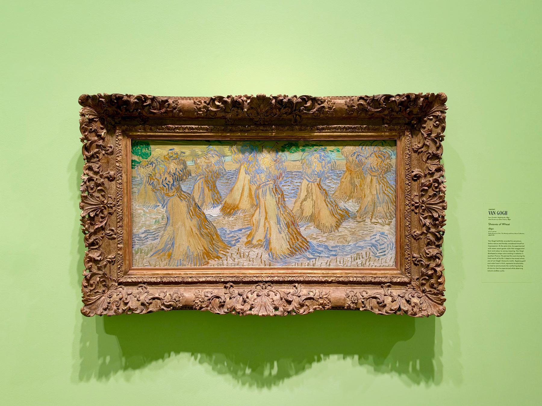 A framed painting hangs on a light green wall. The ornate, carved wooden frame is dark gold-brown with intricate floral and vine patterns along its edges. Inside the frame, the painting depicts golden-yellow sheaves of wheat arranged in multiple stacks across a field, with swirling blue and white brushstrokes suggesting water or sky, characteristic of the artist’s expressive style. Slight patches of green appear in the background, possibly indicating distant trees or foliage. To the right of the frame, a small museum label identifies the artist as Van Gogh and the painting as "Sheaves of Wheat," dated 1890, and provides a brief descriptive text about the artwork. The painting casts a soft shadow onto the wall beneath the frame.