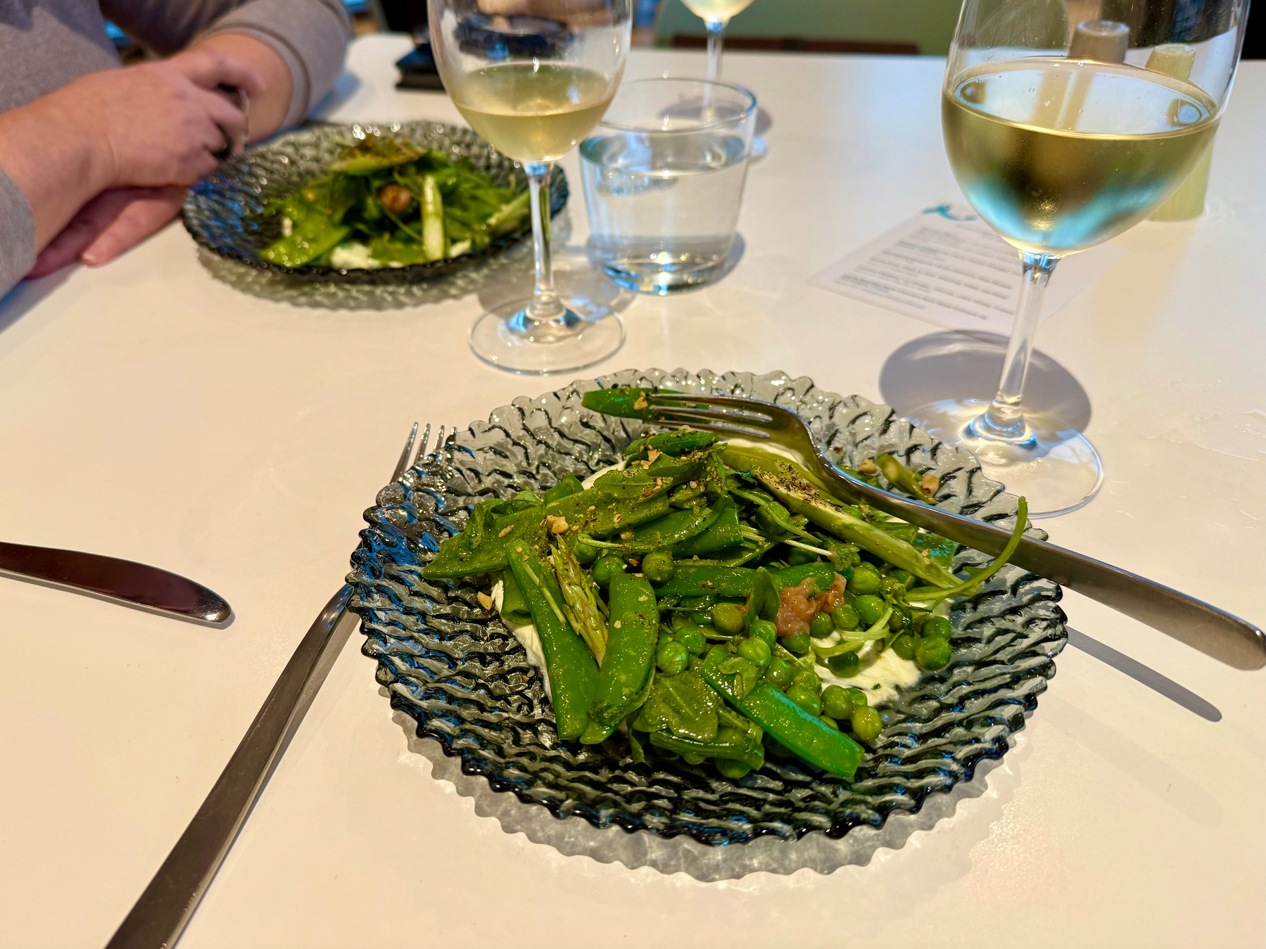 A close-up of a dining table with two elegant, textured glass plates holding fresh green salads made of snap peas, asparagus, and other green vegetables, garnished with seasoning. A fork rests on the plate in the foreground. Each place setting includes a knife and a fork, and two glasses of chilled white wine sit nearby, one in front of each diner. There is also a clear glass of water and a small card on the table. A person with folded arms is seated across the table, wearing a light grey long-sleeve top. The surface of the table is white and clean, and the scene is well-lit with natural light.