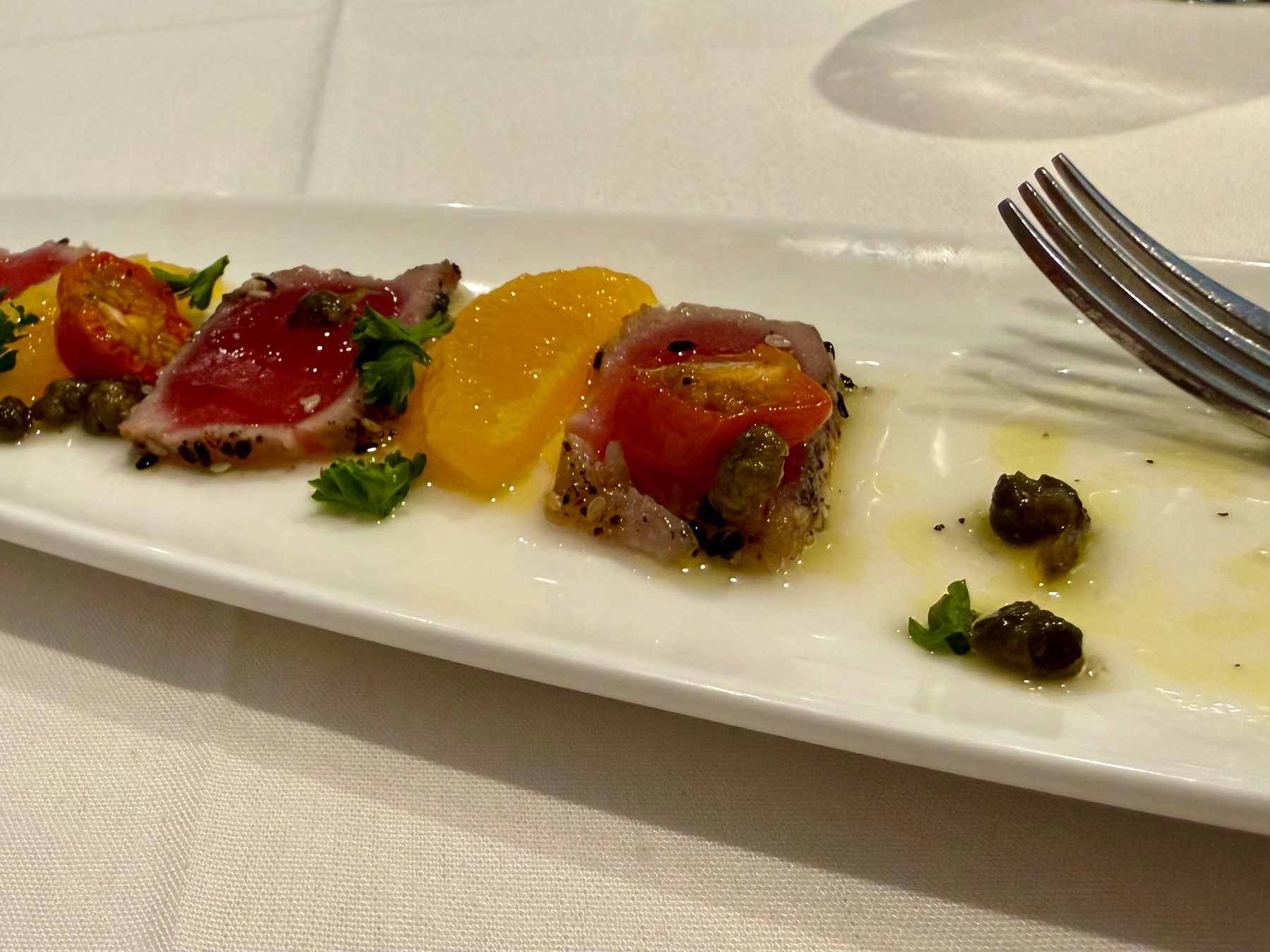A rectangular white plate displays an elegant appetizer consisting of seared tuna slices with a pink centre, garnished with black sesame seeds. The tuna is arranged with orange slices, halved roasted cherry tomatoes, capers, and small sprigs of parsley. A light drizzle of olive oil is visible on the plate, along with a few scattered capers and herbs near the right side. A fork rests at the edge of the plate, and the background features a white tablecloth with a faint shadow of a glass.