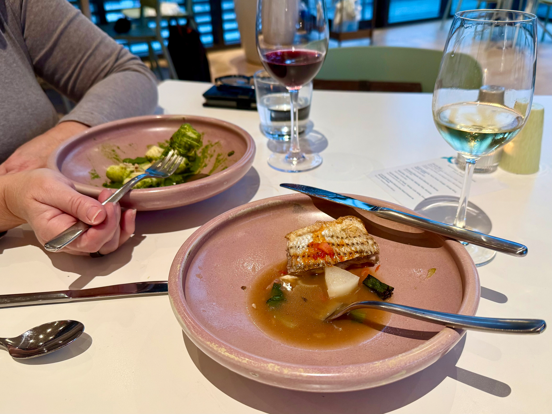Two partially eaten dishes of food are on a white table in a modern dining setting. The foreground plate has a piece of grilled fish with crispy skin, a chunk of white root vegetable, and a few vegetables in a light brown broth. A fork and knife rest on the edge of the plate. The background plate has green gnocchi or vegetables coated in a green sauce, with a hand holding a fork spearing a piece. A glass of red wine, a glass of white wine with condensation, and a small water glass are also on the table. The background shows a bright, contemporary dining area with glass walls and metal chairs.