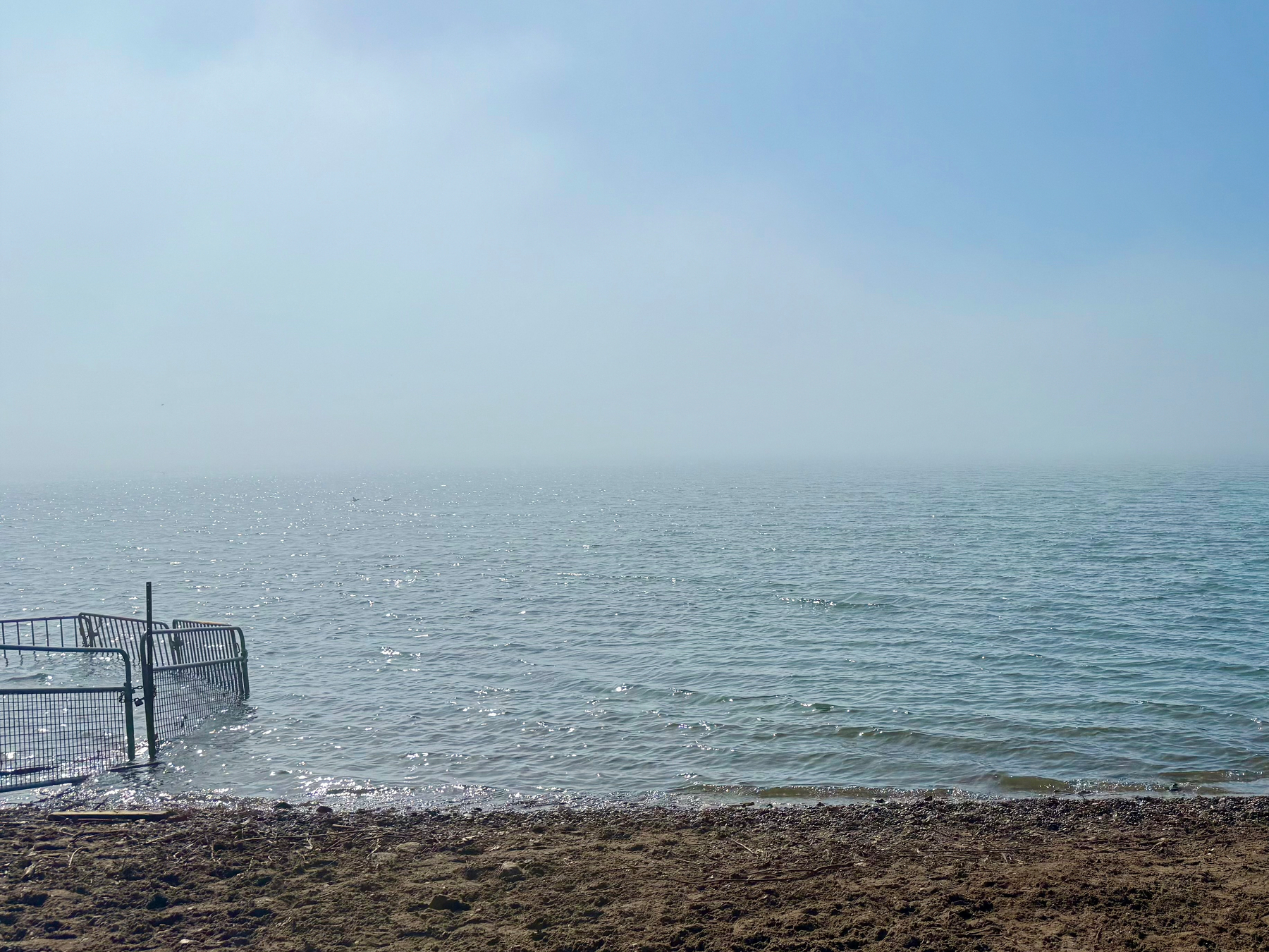 A misty lakeside scene with calm, rippling water stretching to the horizon under a hazy blue sky. The far distance fades into a soft fog, blending lake and sky almost seamlessly. In the foreground, there is a sandy and slightly rocky shoreline. To the left, a partially submerged metal fence extends from the shore into the water, with its lower section surrounded by small waves that glint in the sunlight.