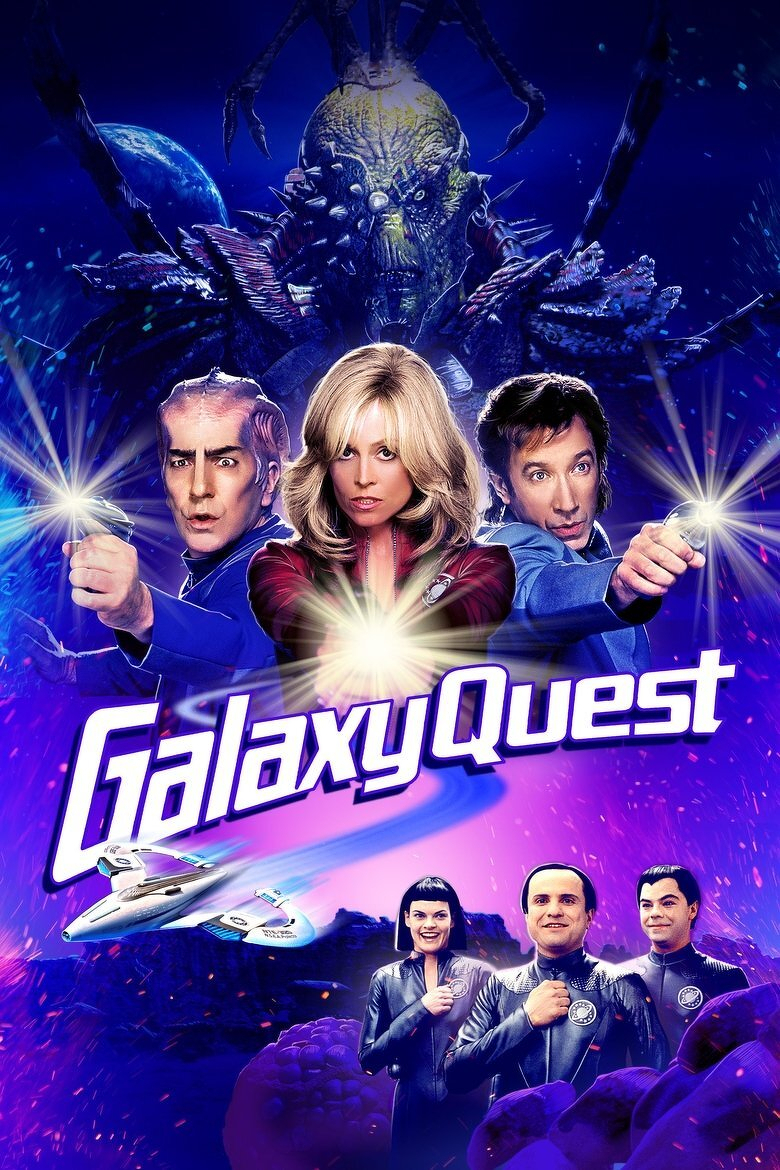 A vibrant sci-fi movie poster features three main characters in the foreground, holding futuristic laser guns pointed outward. The man on the left has slicked-back hair and a serious expression, the woman in the centre has blonde hair with bangs and a determined look, and the man on the right has dark hair and is aiming his weapon forward. Behind them, a large menacing alien creature with insect-like features looms against a backdrop of stars and a distant planet. Below the three characters, a sleek white spaceship with blue accents flies through space. Standing proudly at the bottom of the image are three additional characters in dark jumpsuits with emblems on their chests, smiling and holding their right fists to their hearts. The title “Galaxy Quest” is prominently displayed in bold, stylized, purple and white letters across the middle of the poster. The overall colour scheme is dominated by deep blues, purples, and bright flashes of light, emphasizing a cosmic adventure theme.