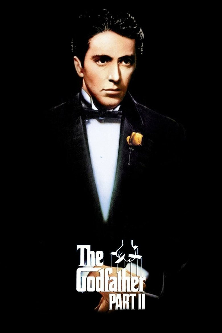 A formal portrait of a man wearing a black tuxedo with a white shirt, black bow tie, and a yellow rose pinned to his lapel, set against a completely black background. He has dark, slicked-back hair and a serious expression, looking slightly to the side. Near the bottom of the image, the text reads “The Godfather Part II” in white, with a puppet hand logo above the word “Godfather.”