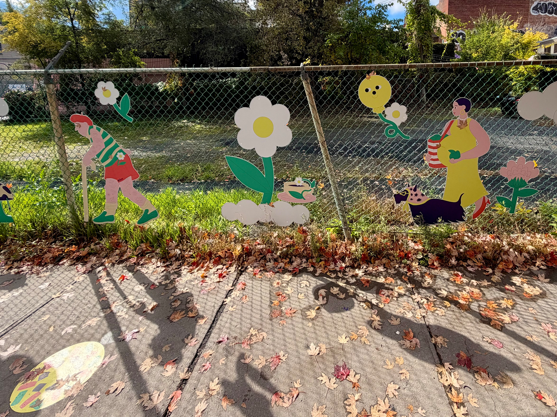 A chain-link fence decorated with colorful cutouts: a person in a red hat and green striped shirt walking with a cane, a large white flower with a yellow center and green leaves, a small figure lying on a white cloud, a yellow round character holding a white flower, a person in a yellow dress holding a red-and-white striped object, a black cat with a red collar, and a pink flower. The fence is in front of grass and trees, with fallen leaves on the sidewalk below. A yellow oval with a green pattern lies on the ground. In the upper right, a sign shows the numbers 4951.
