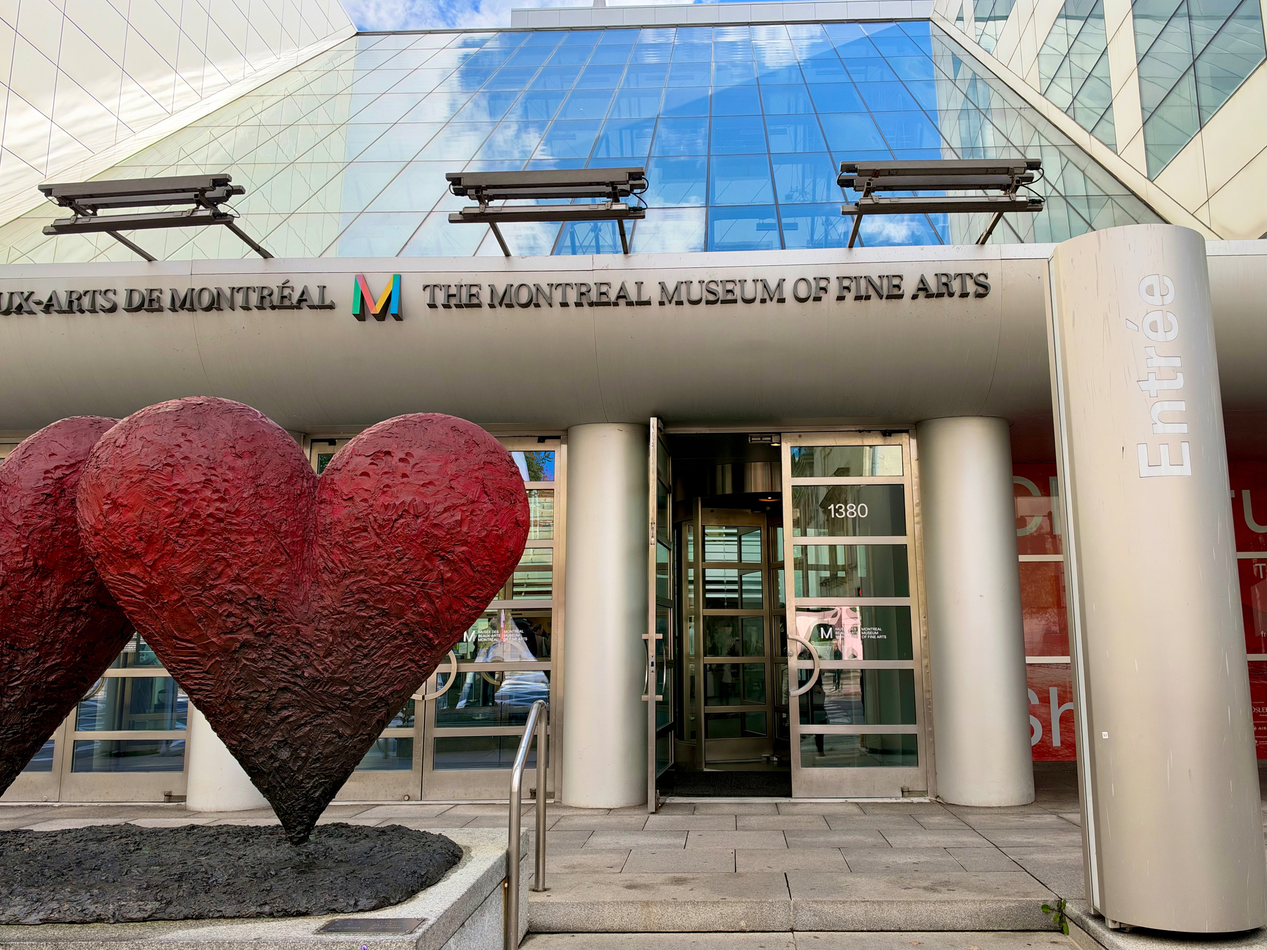 Two large textured red heart sculptures in a dark base stand before a glass-fronted building with a sloped glass roof reflecting clouds. Above the entrance, black text reads “UX-ARTS DE MONTRÉAL” beside a multicolored M logo and “THE MONTREAL MUSEUM OF FINE ARTS.” A vertical pillar on the right shows white text “Entrée.” The glass door displays the number “1380.” Two metal structures extend above the entrance, and a metal railing leads to the doorway.