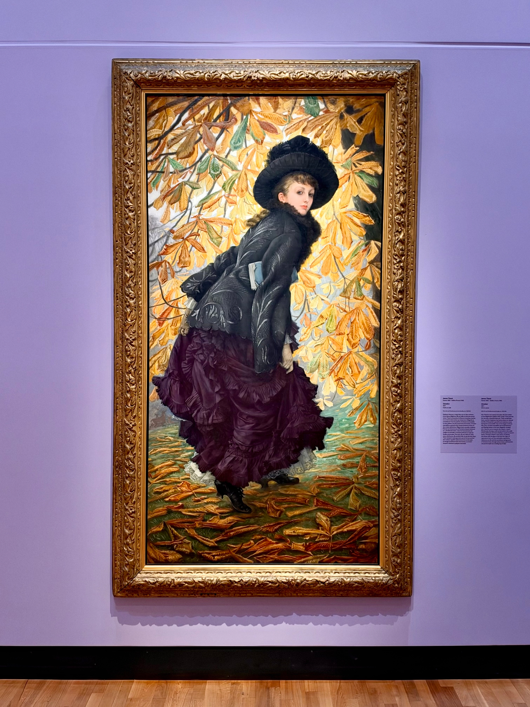 A vertical painting in an ornate gold frame on a lavender wall shows a woman in a wide black hat and dark patterned coat with a ruffled purple skirt, standing among yellow and orange leaves on the ground and in the background. She looks back over her shoulder. A small rectangular label with text is mounted to the right of the frame.