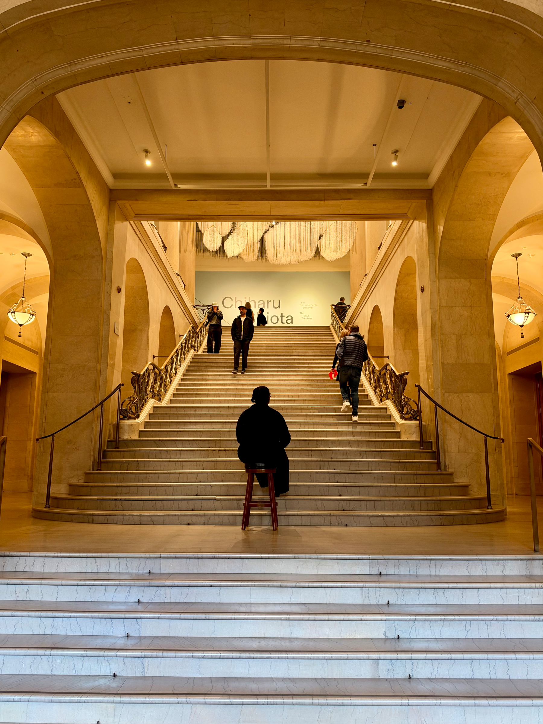 A person in dark clothing sits on a red stool at the base of a wide marble staircase in a grand hall with arched stone walls and ornate railings. Several people ascend the stairs toward a wall displaying the text “Chiharu Shiota.” A large hanging installation of pale threads or cords is suspended above the text. Warm light from hanging lamps and ceiling fixtures illuminates the scene.