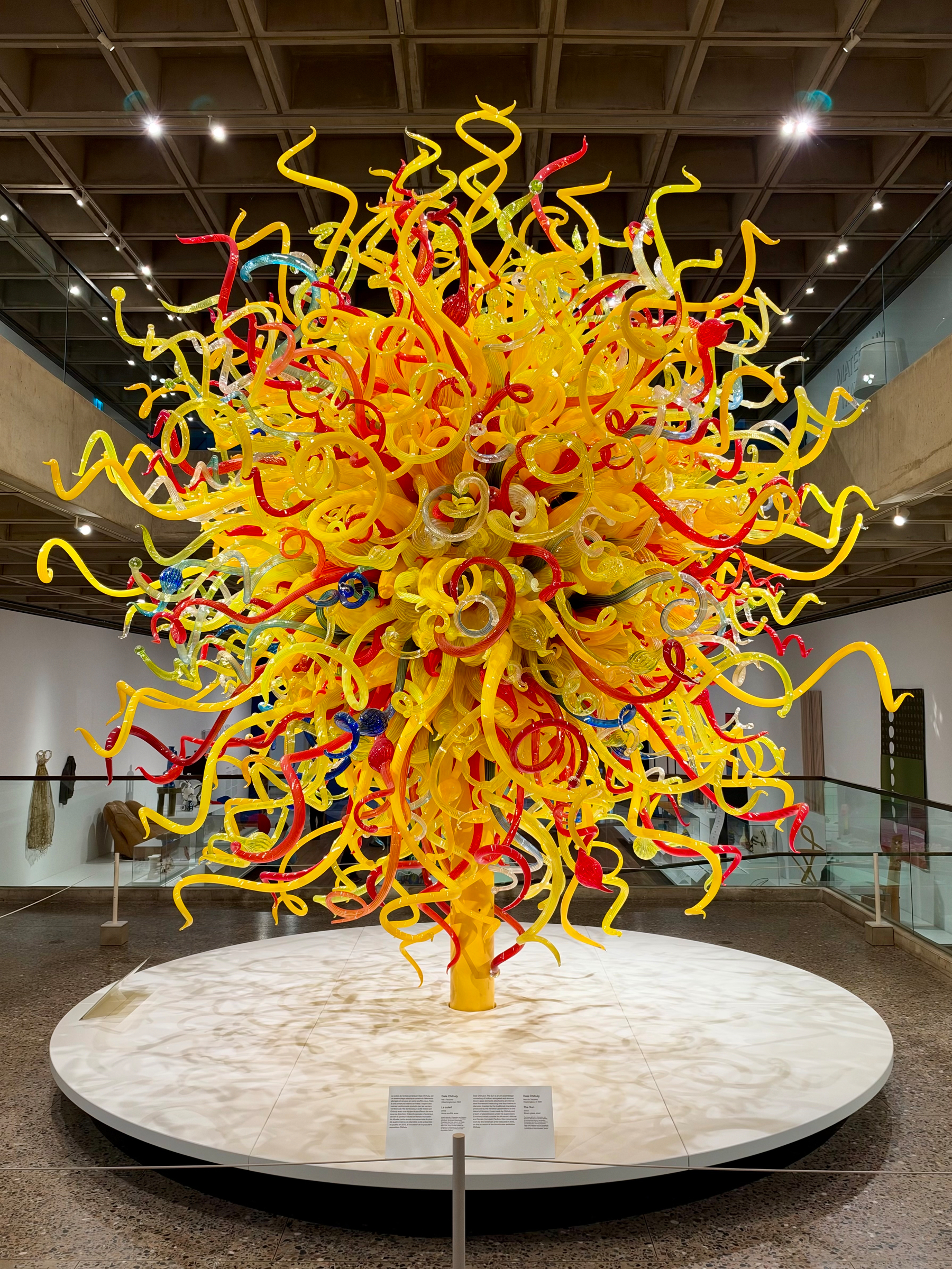 A large sculpture of twisting yellow, red, blue, and clear glass tendrils rising from a yellow base, resembling a burst, centered on a circular white platform in a gallery with a coffered ceiling and glass railings. A small rectangular plaque with multiple lines of text sits on the platform in front.
