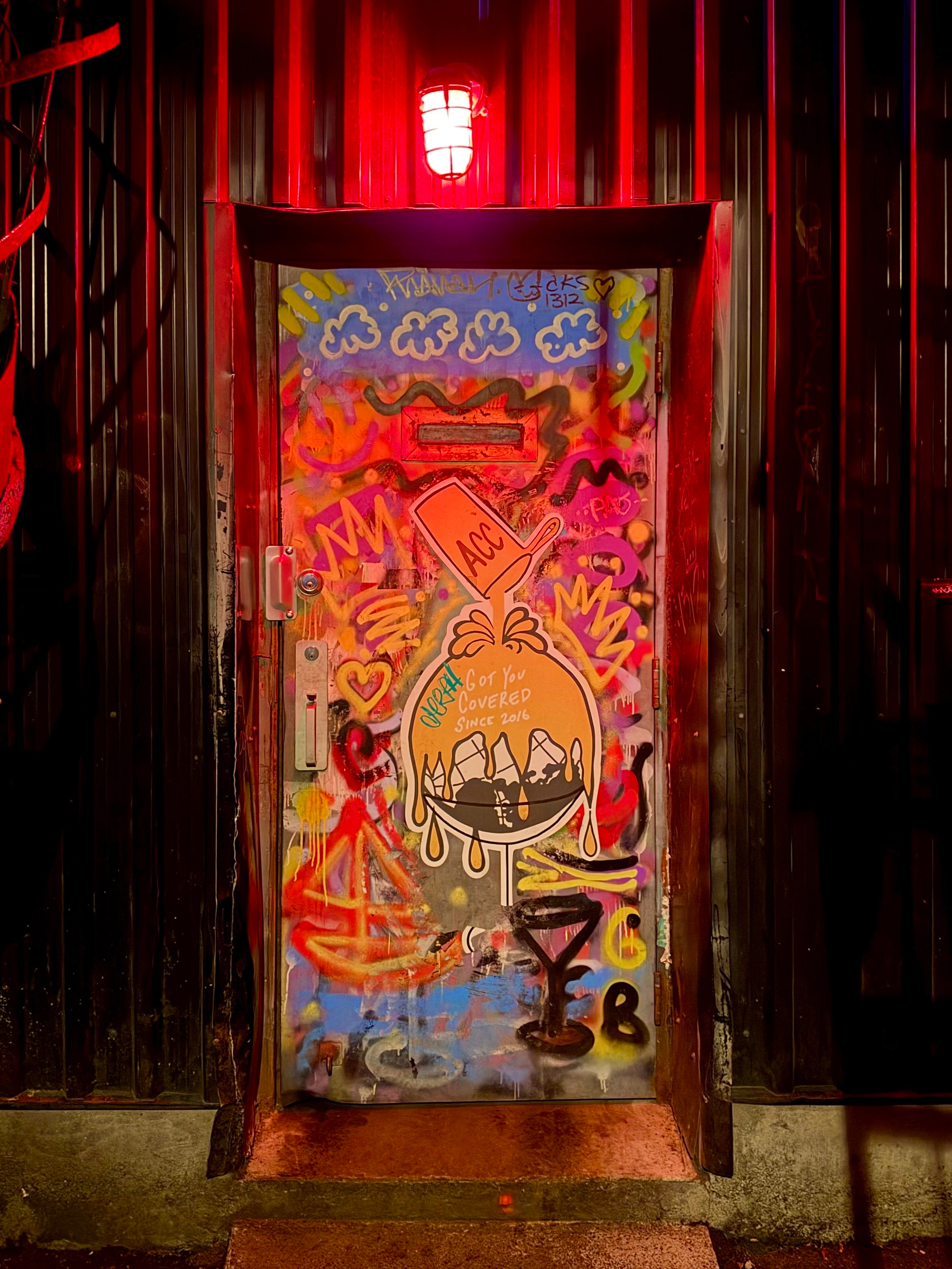 A graffiti-covered door under a glowing red light in a dark corridor with corrugated walls. The door features colorful spray paint including four white clouds at the top, a dripping orange bag with a bowl holding three white shapes, and the text “Got You Covered Since 2016” inside the bag. Above it, a tilted orange shape contains “ACC”. Other markings include “JCS 132”, “NHA”, “M”, a heart, and abstract shapes in red, yellow, blue, and black. A silver handle and lock are on the left. The door sits on a worn step with a dark frame and red reflections.