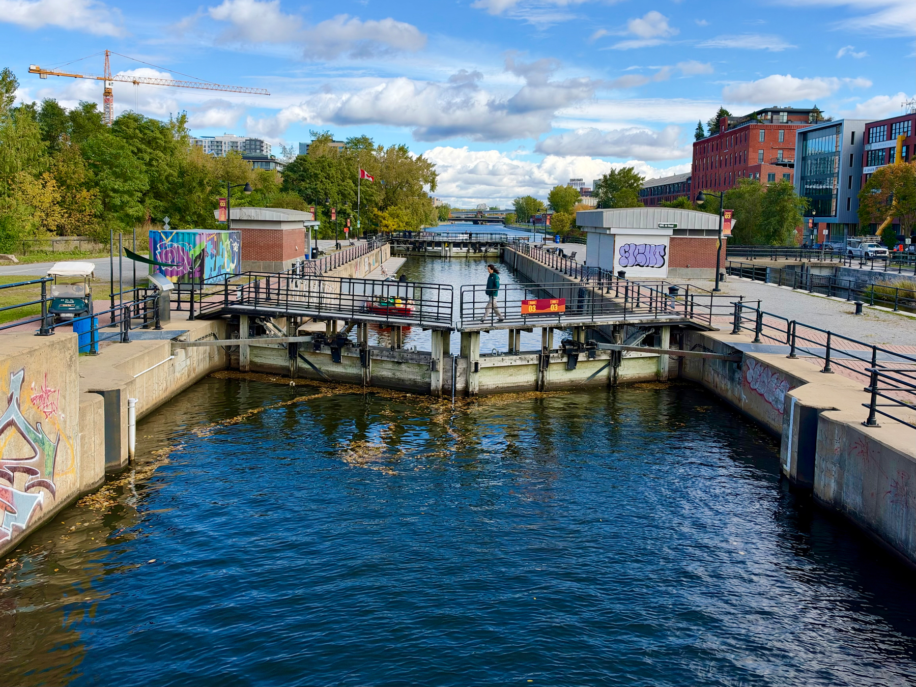 A canal lock with dark blue water flowing between concrete walls lined with black railings. A person stands on a metal walkway crossing the lock, near two small brick buildings. Colorful graffiti appears on the left wall, the right wall, and both brick buildings. Trees and buildings, including a red brick structure and a modern glass building, line the background under a blue sky with white clouds. A construction crane rises above the trees on the left.