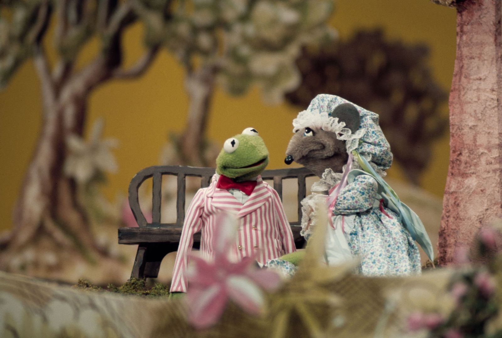 A scene from The Muppet Show featuring Kermit the Frog and a female mouse puppet dressed in a blue floral dress with a matching bonnet. Kermit is wearing a pink and white striped suit jacket with a red bow tie. They are sitting together on a wooden bench in front of a backdrop with stylized trees and a yellow background. Pink and cream flowers are blurred in the foreground, adding depth to the whimsical outdoor setting.