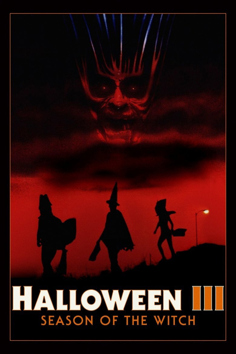 A movie poster with a dark red and black color scheme. Across the upper half, a huge, grimacing face with hollow, glowing red eyes and a wide, distorted mouth fills the smoky clouds. Thin streaks of blue and white light radiate upward from its head like wild hair. Below, three black silhouettes of trick-or-treaters stand on a grassy hill against a blood-red sky. The figure on the left wears a cape and carries a bulky bag, the one in the center wears a tall pointed witch's hat and a flowing cloak and holds a long stick, and the one on the right wears a hat with pointed ears and carries a bucket. A glowing streetlamp stands on the right edge. A thin orange border frames the poster. At the bottom, large white capital letters read "HALLOWEEN" with a stylized orange "III" beside it, and smaller orange capital letters below read "SEASON OF THE WITCH."