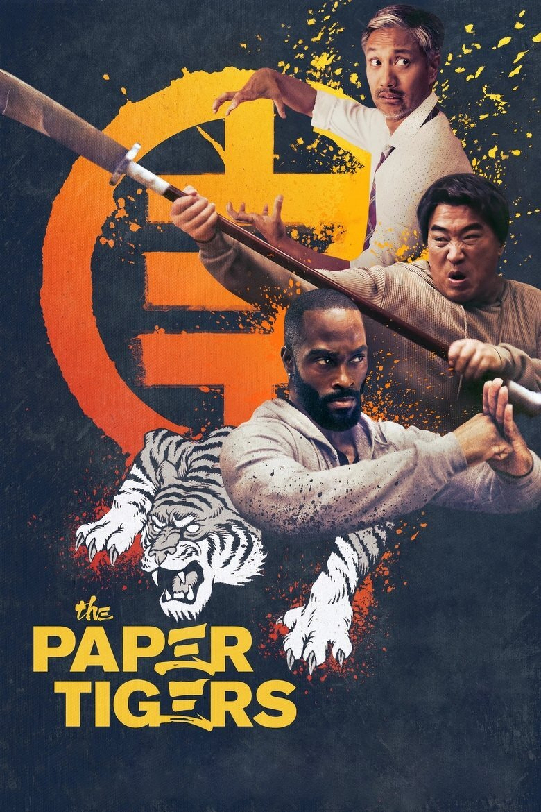Three men in martial arts poses are featured against a dark, textured background with splashes of orange and yellow paint. The man at the top right, with short grey hair and a white dress shirt with a tie, looks over his shoulder in a defensive stance. Below him, another man with medium-length black hair and a beige hoodie aggressively wields a wooden staff, his face contorted in a shout. At the bottom, a bearded man in a grey hoodie is posed in a strong kung fu stance, his fists clenched and eyes focused. Behind them is a large orange circle with a stylized Chinese character in the centre, and a white tiger snarling and leaping forward from the bottom left. Yellow text at the bottom reads “the PAPER TIGERS,” with the word “PAPER” featuring claw marks and stylized lettering.