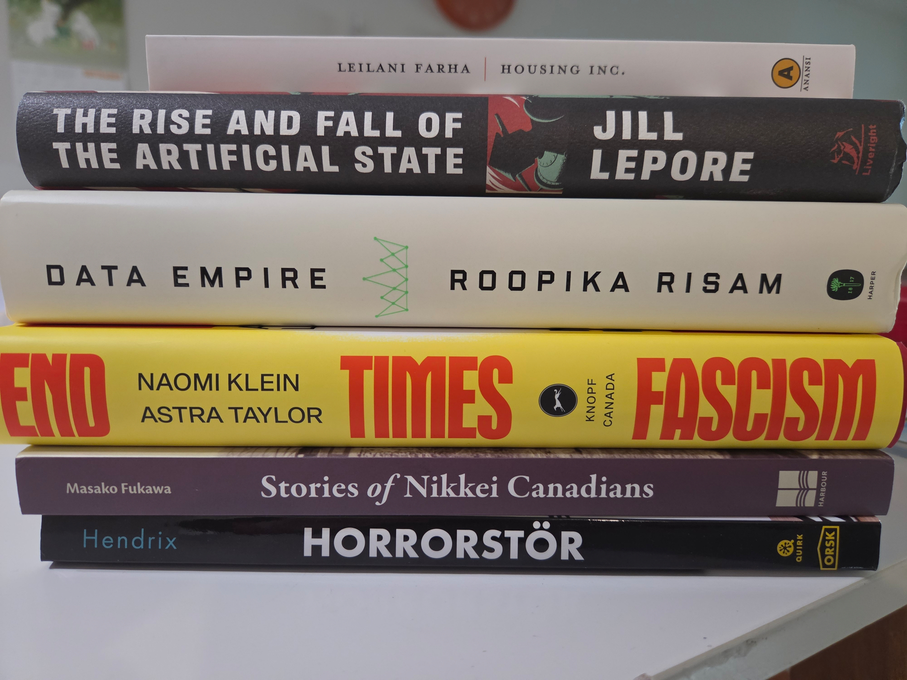 Photo of a stack of books. From bottom to top the titles are: Horrorstör, Stories of Nikkei Canadians, End Times Fascism, Data Empire, The Rise and Fall of the Artificial State, Housing Inc.