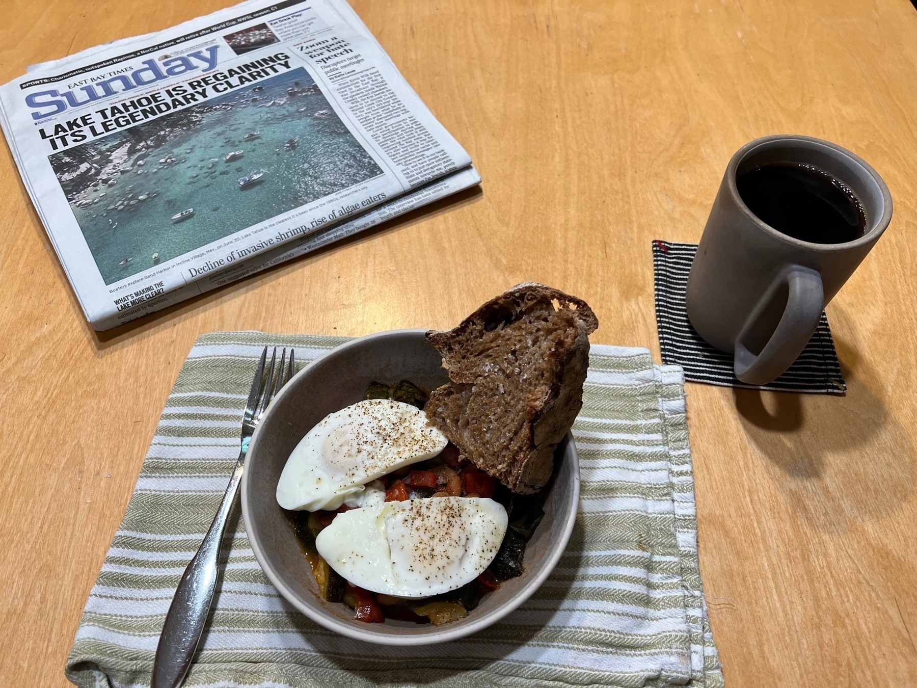 A bowl with poached eggs on top of garbanzo beans, kale, and tomatoes. A cup of coffee and a newspaper. 