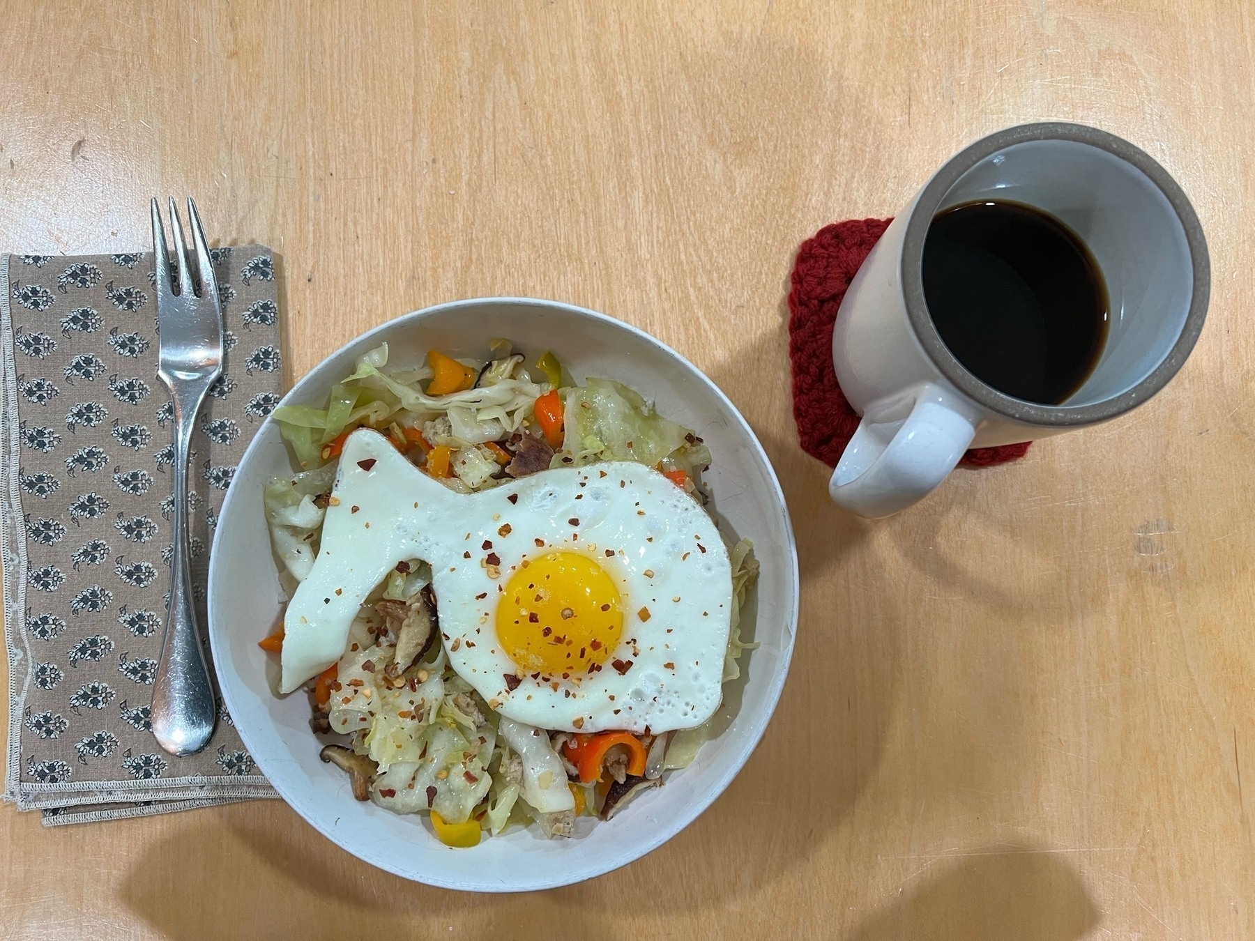 Top down view of a bowl of sautéed cabbage, peppers, and a sunny side up egg. Coffee in a white ceramic cup, a fork on a cloth napkin. 