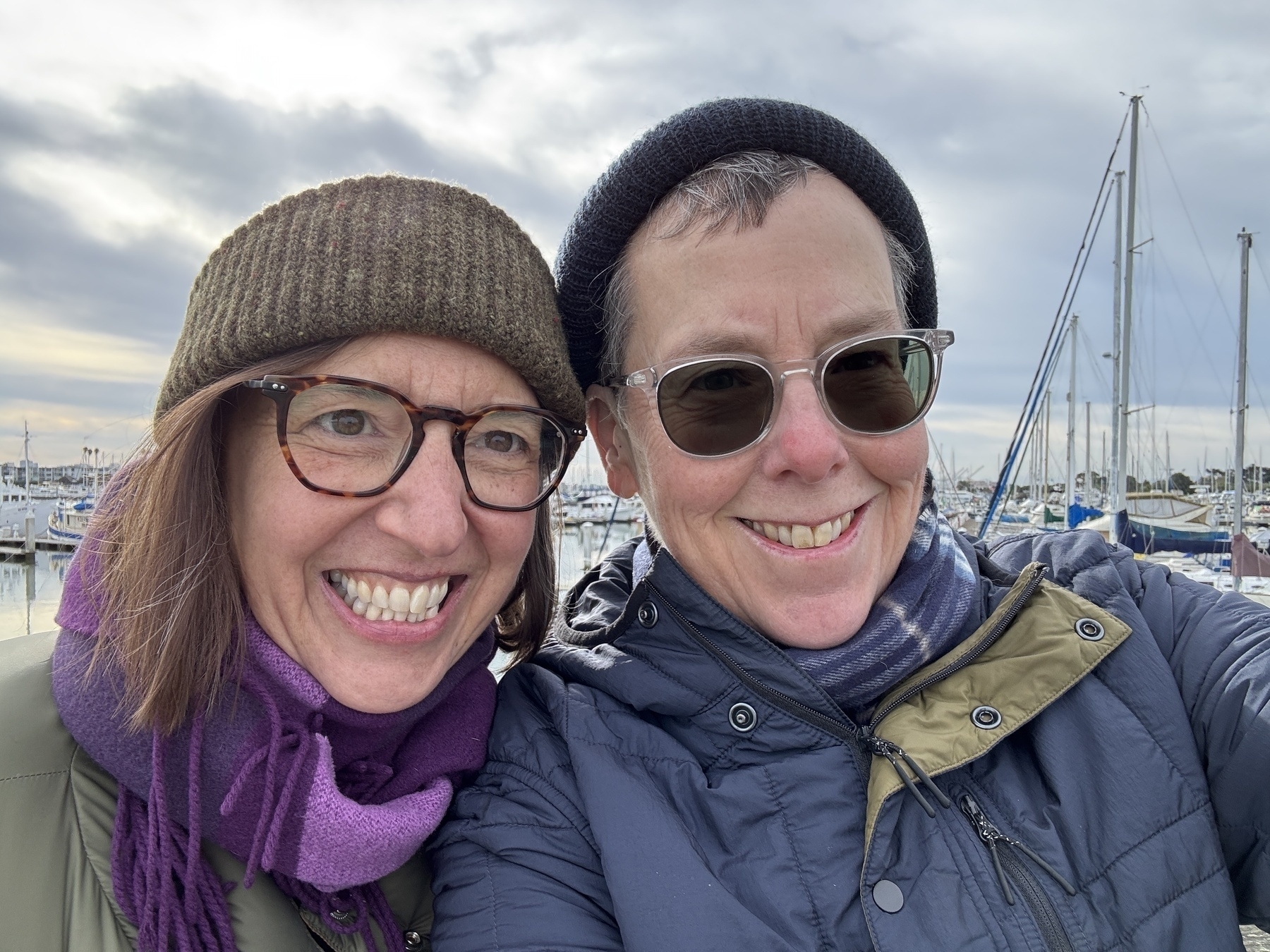 Two people, in beanies, scarves, and warm jackets smiling at the camera. Boats in the background. 