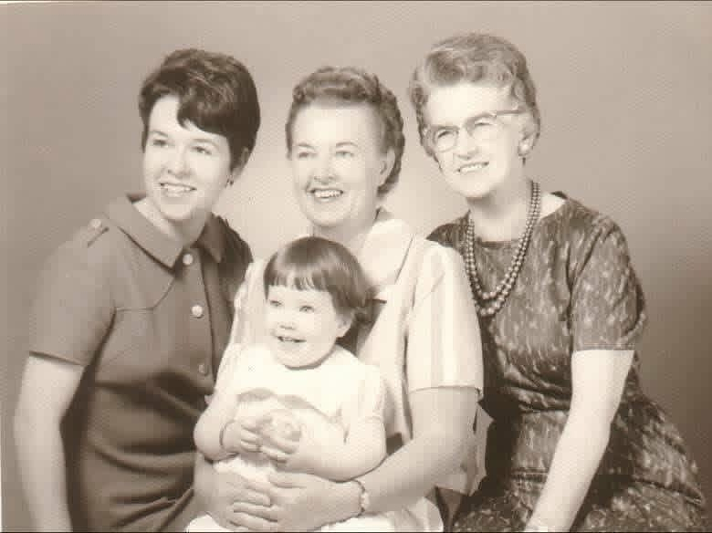 Four generations of smiling women, ranging from a young girl to elderly, pose together in a black and white photo.