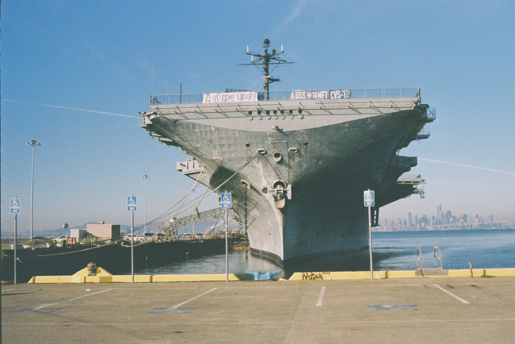 An aircraft carrier is docked at a harbor near an empty parking lot, with a city skyline visible in the background.