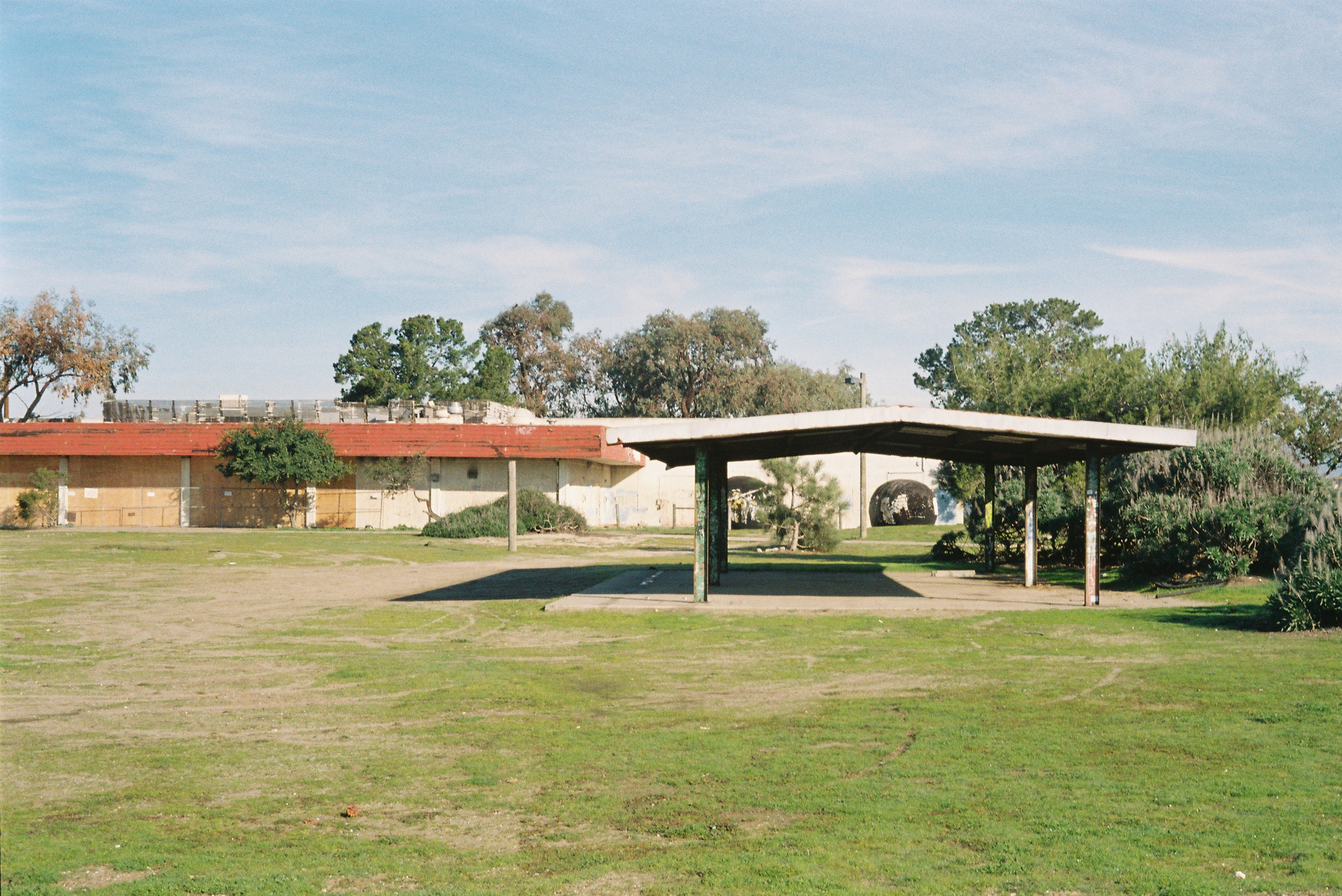 A pavilion stands in a grassy open area with a building and trees in the background.