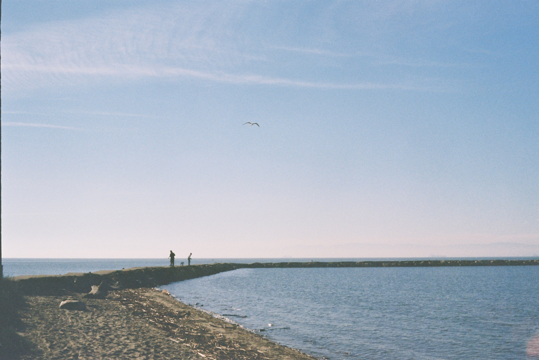 A peaceful coastal scene shows two people standing on a narrow jetty extending into the sea under a clear blue sky.