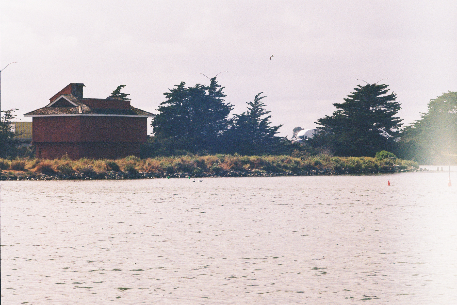 A red house and trees are situated by the edge of a body of water under a cloudy sky.