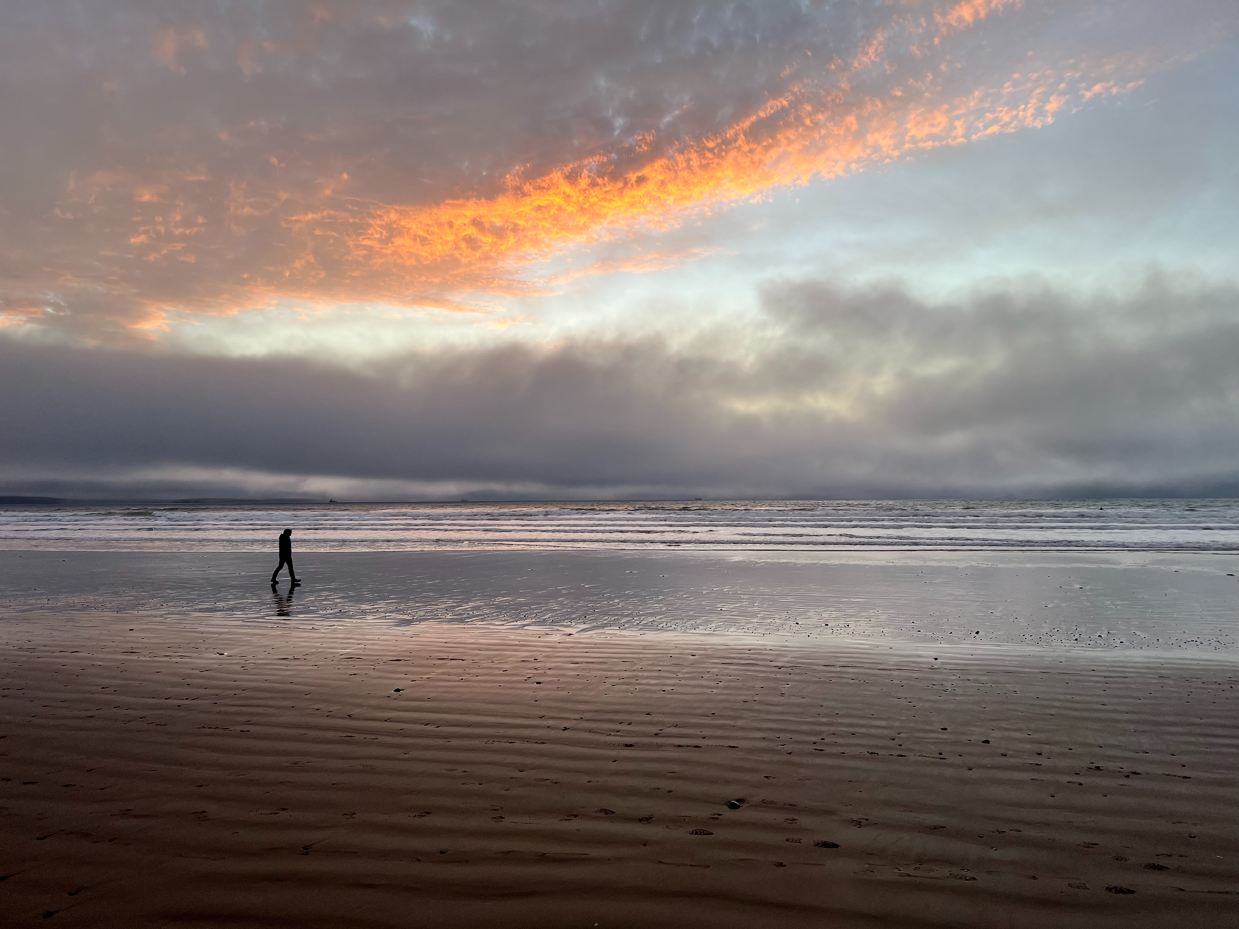 A person walks along a vast, reflective beach under a dramatic, colorful sky.