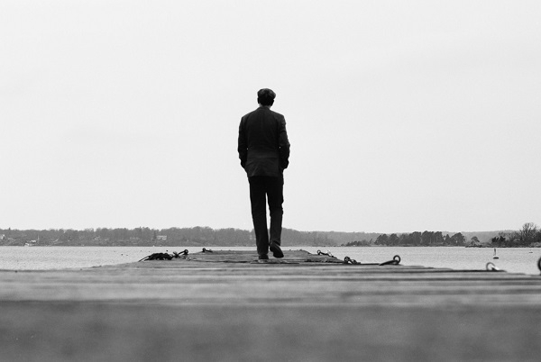 A black and white photo of a man walking away from the camera out on to a jetty.