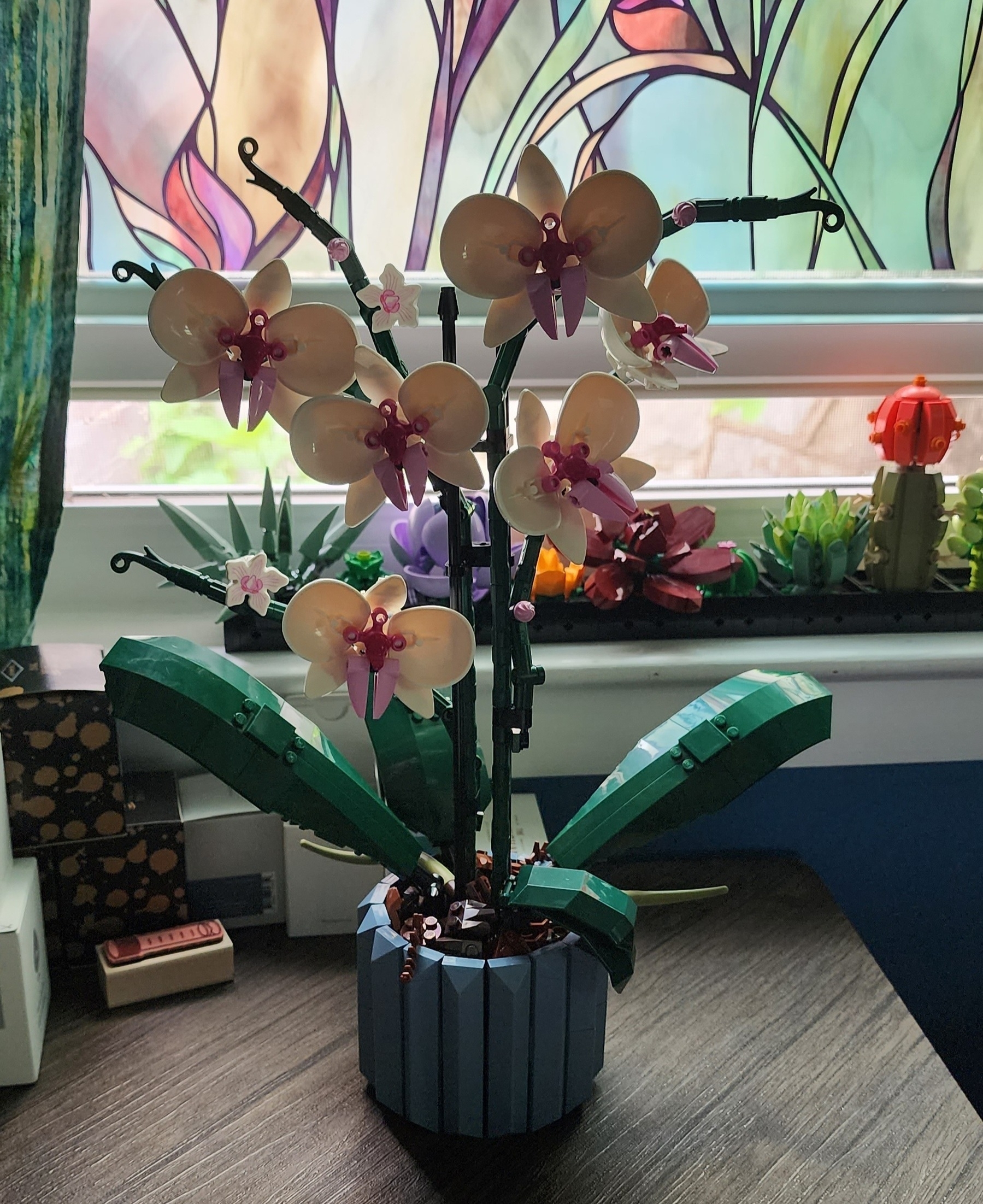 A completed Lego Orchid plant build with six large white and pink blooms, several more buds along the stems, and large dark green leaves, all set in a smal blue-gray Lego planter with brown pieces representing dirt and mulch.