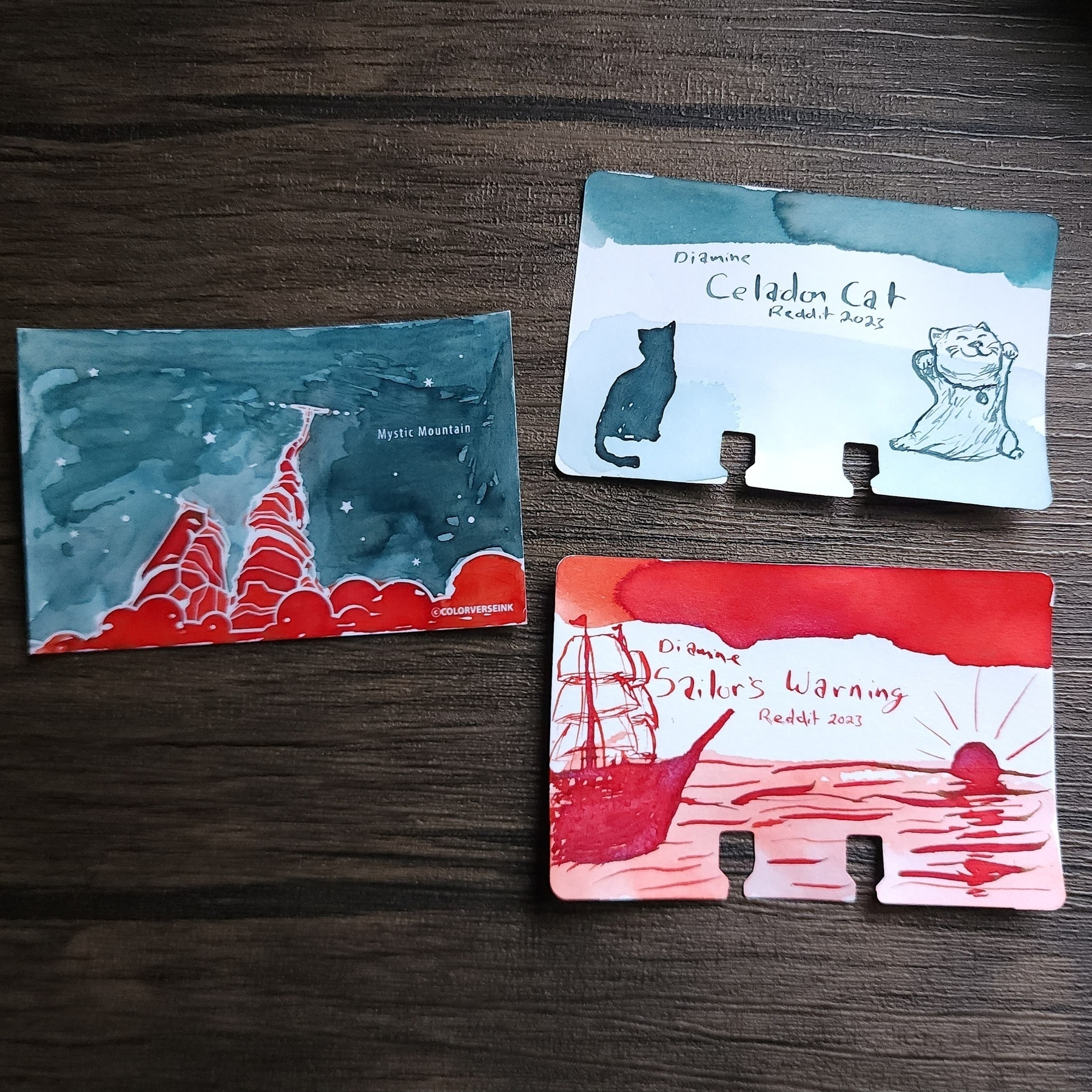 Ink swatch cards for Diamine Celadon Cat, a medium blue green color, and Sailor's Warning, a bright brick red with pinkish shimmer.