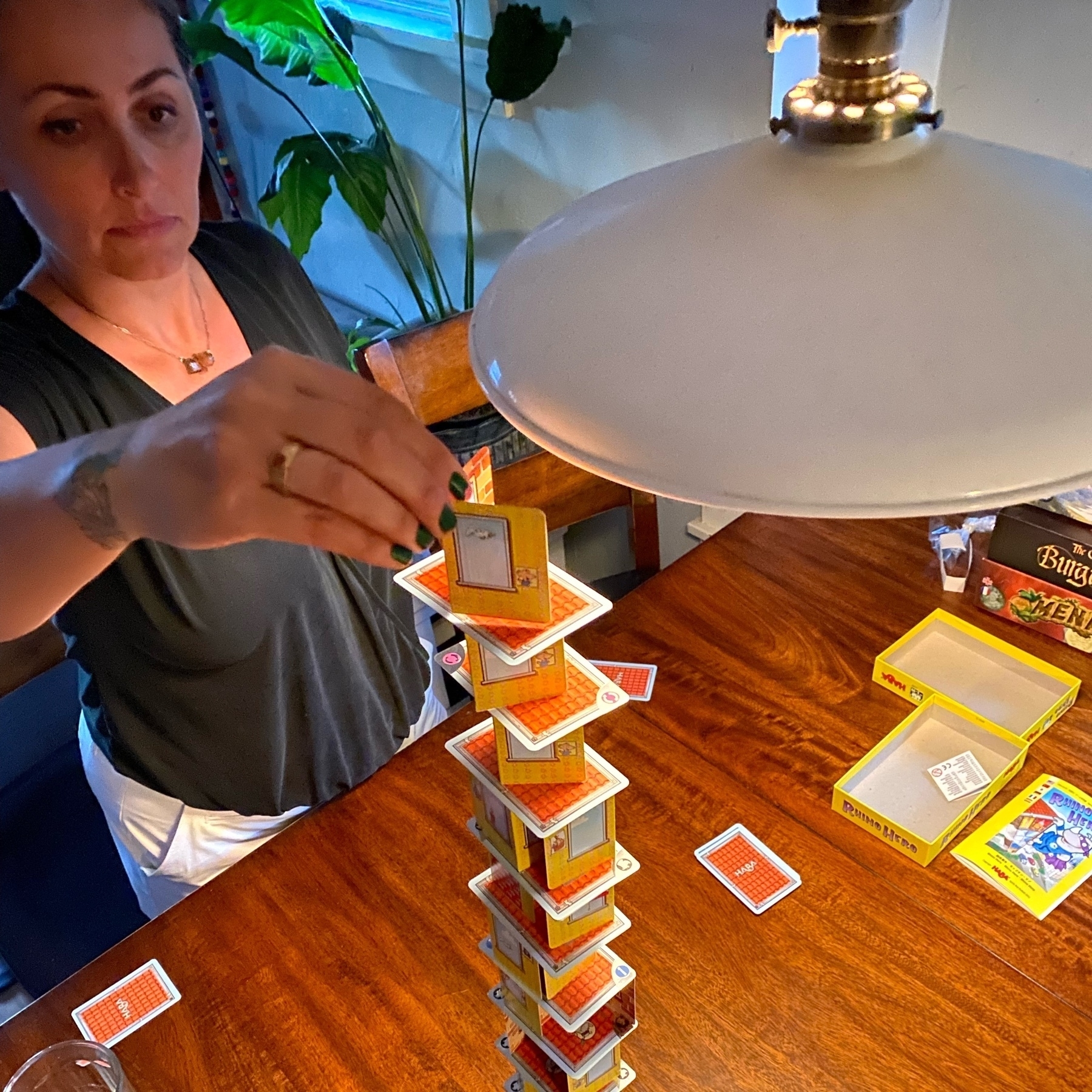 Lauren delicately places a structure card to the Rhino Hero tower. The card tower is so high it is about to hit the hanging lamp. 