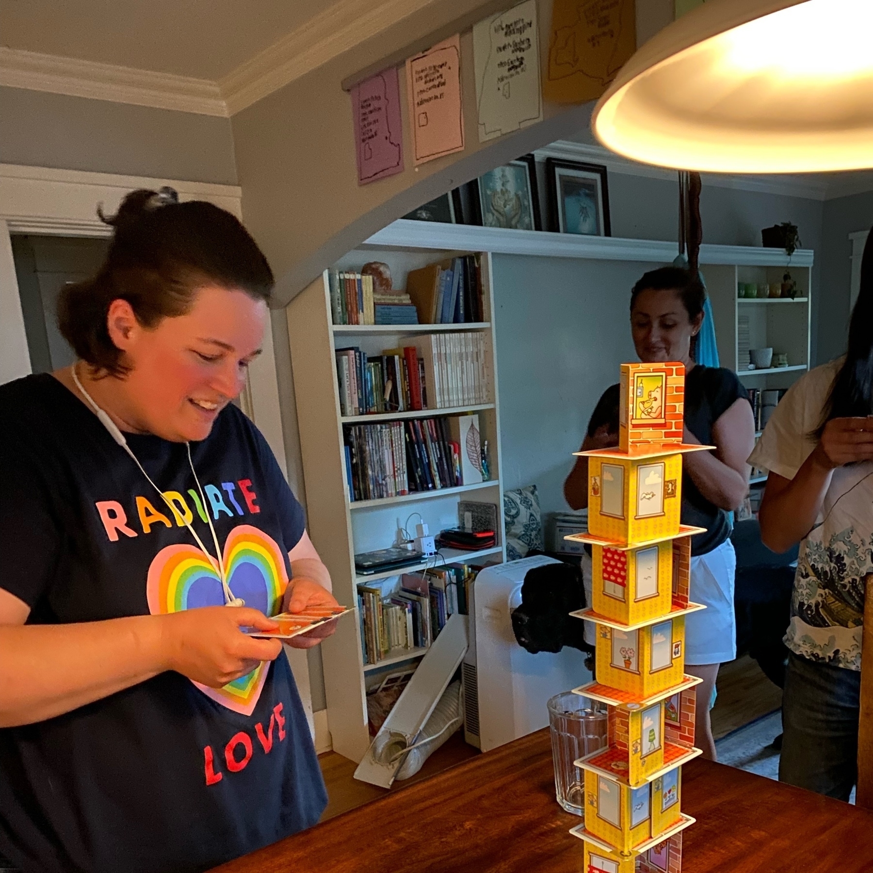 Laura looking at the roof she must build next for Rhino Hero, a stacking game. She is playing in a dining room. The stack is high and sobblomg already. 