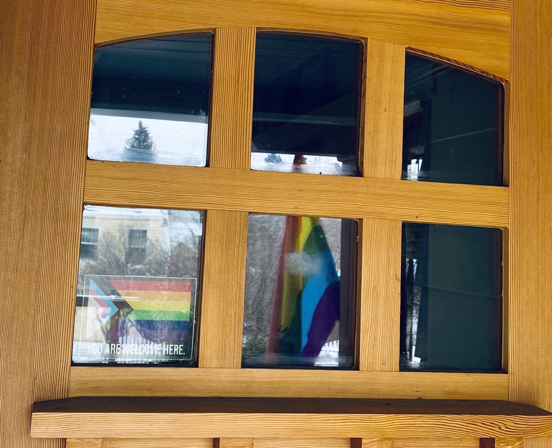 Six glass tiles at the top of a wooden front door. A pride “you are welcome here” is in the bottom left; a pride flag and a winter snowy morning is reflected in the other tiles. 
