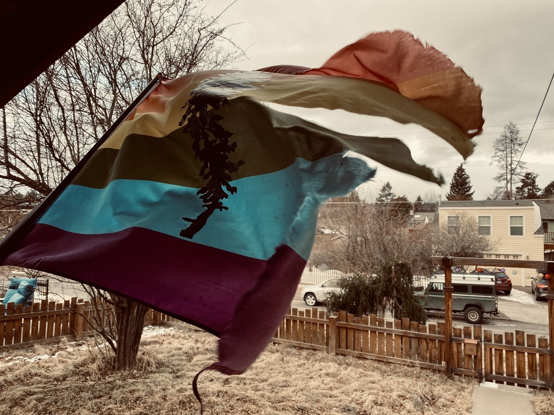 Tattered Pacific Northwest pride flag flapping dramatically in the wind. The flag is on a front porch overlooking a front lawn with a small wooden picket fence. 