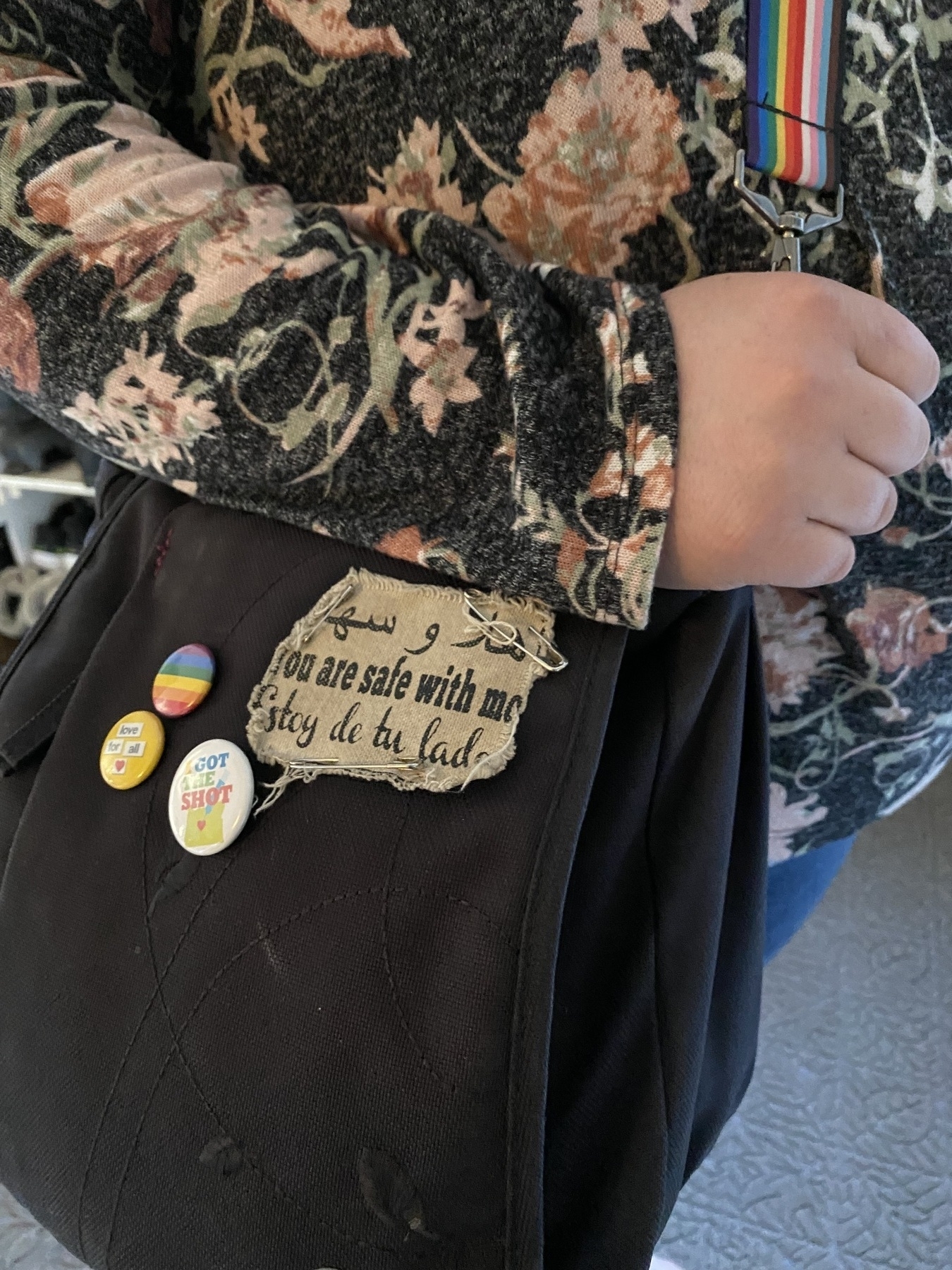 Woman wearing black, cloth purse over shoulder. There is a patch that reads “You are safe with me.” There is also a round pride pin. 