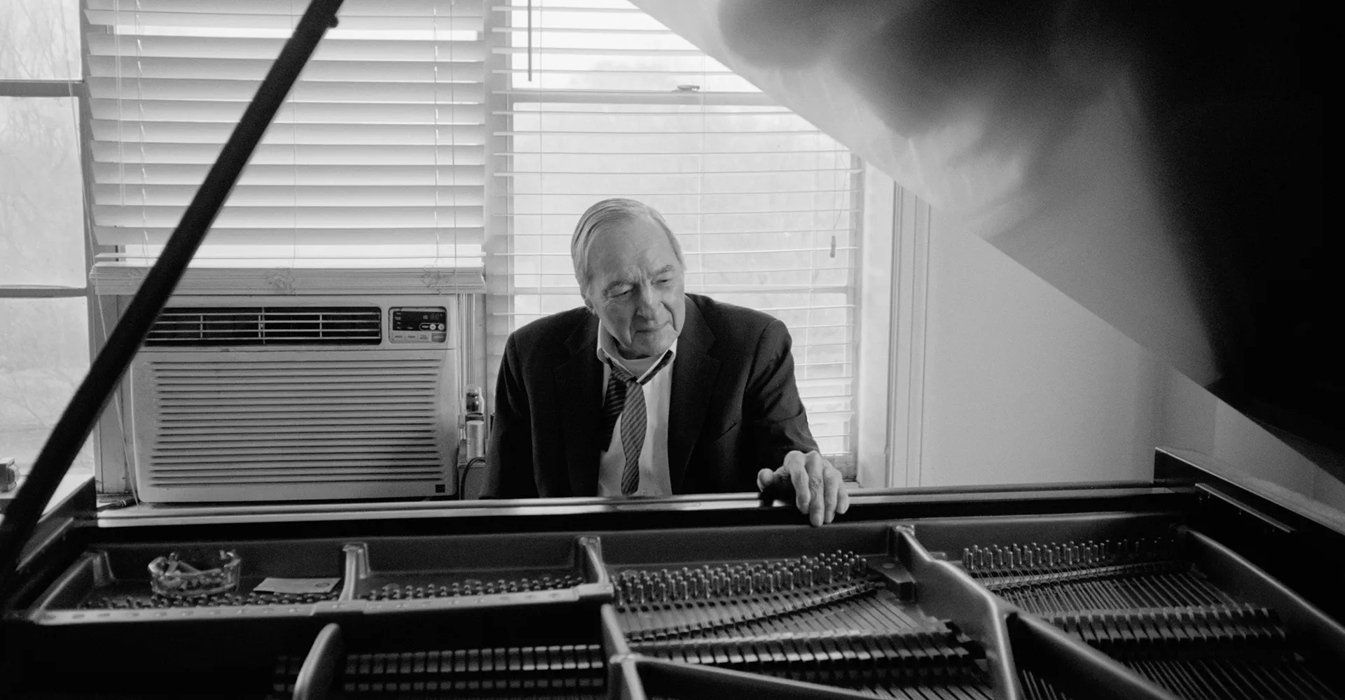 Black and white portrait of photographer William Eggleston at a piano with white metal blinds behind him. His hand is resting just above the hammers in the interior of the baby grand