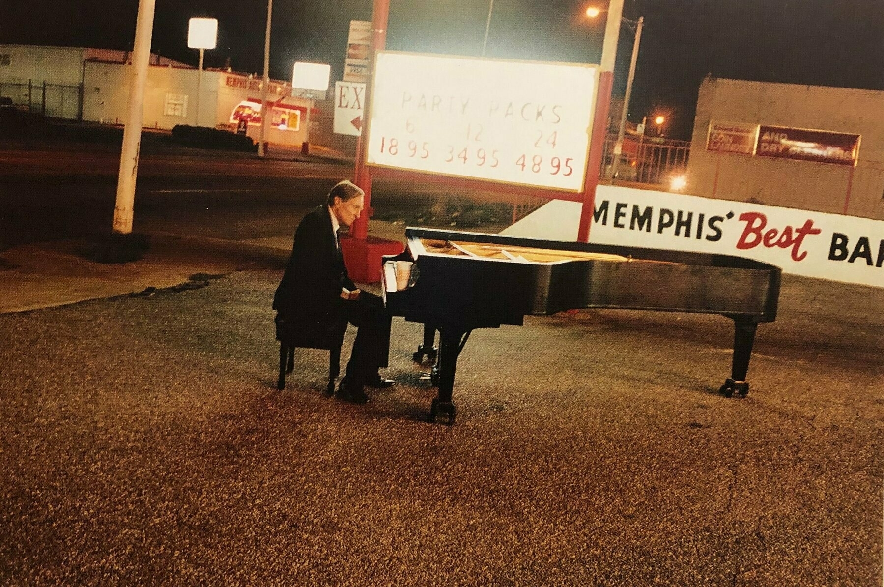 William Eggleston playing a battered baby grand piano at night on a roadside illuminated by a bar's glowing sign