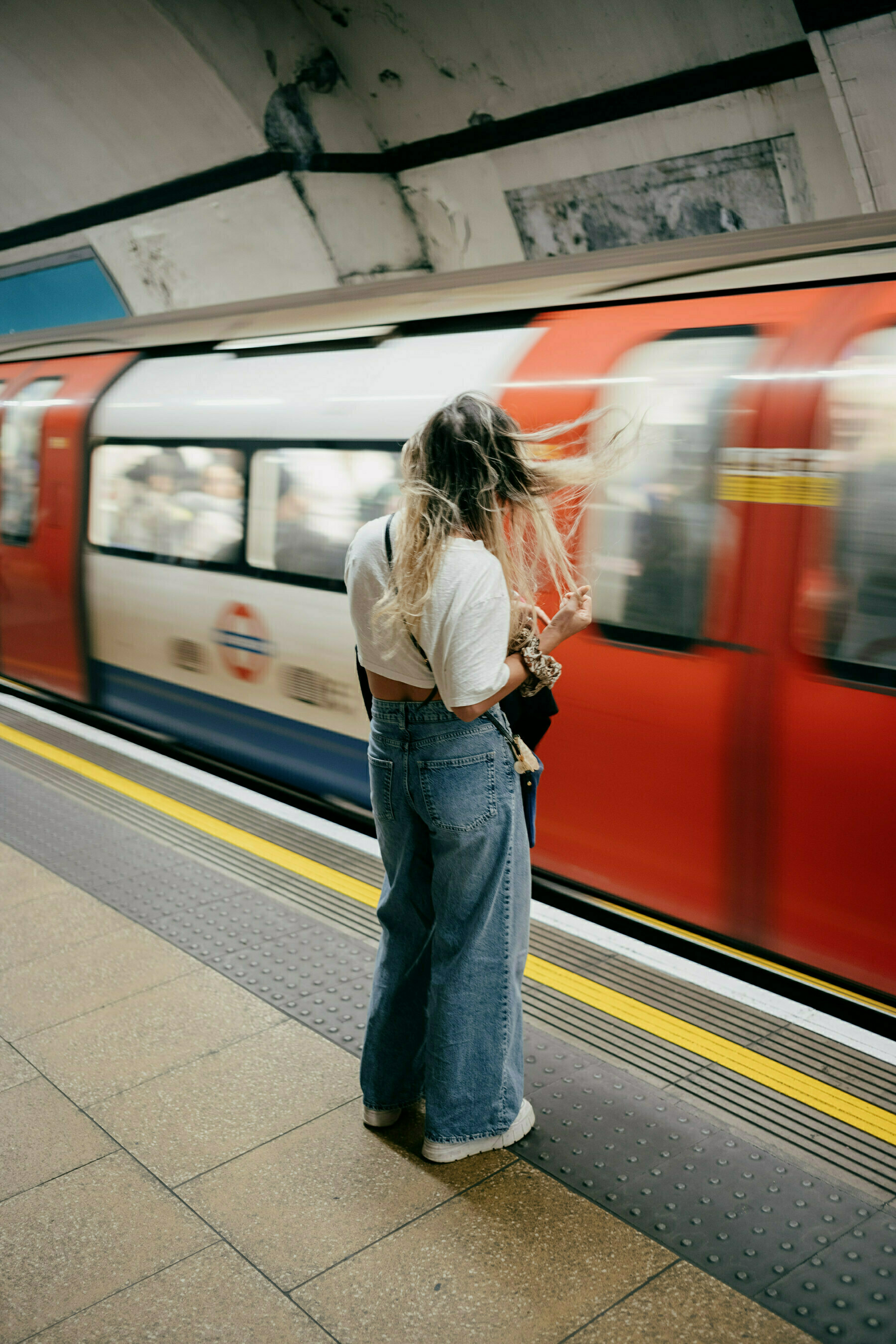 Full length shot of girl wearing blue jeans and a white top standing on a tube platform with her hair blowing in the wind created by the tube train rushing into the station. The tube train is blurred from the movement. 