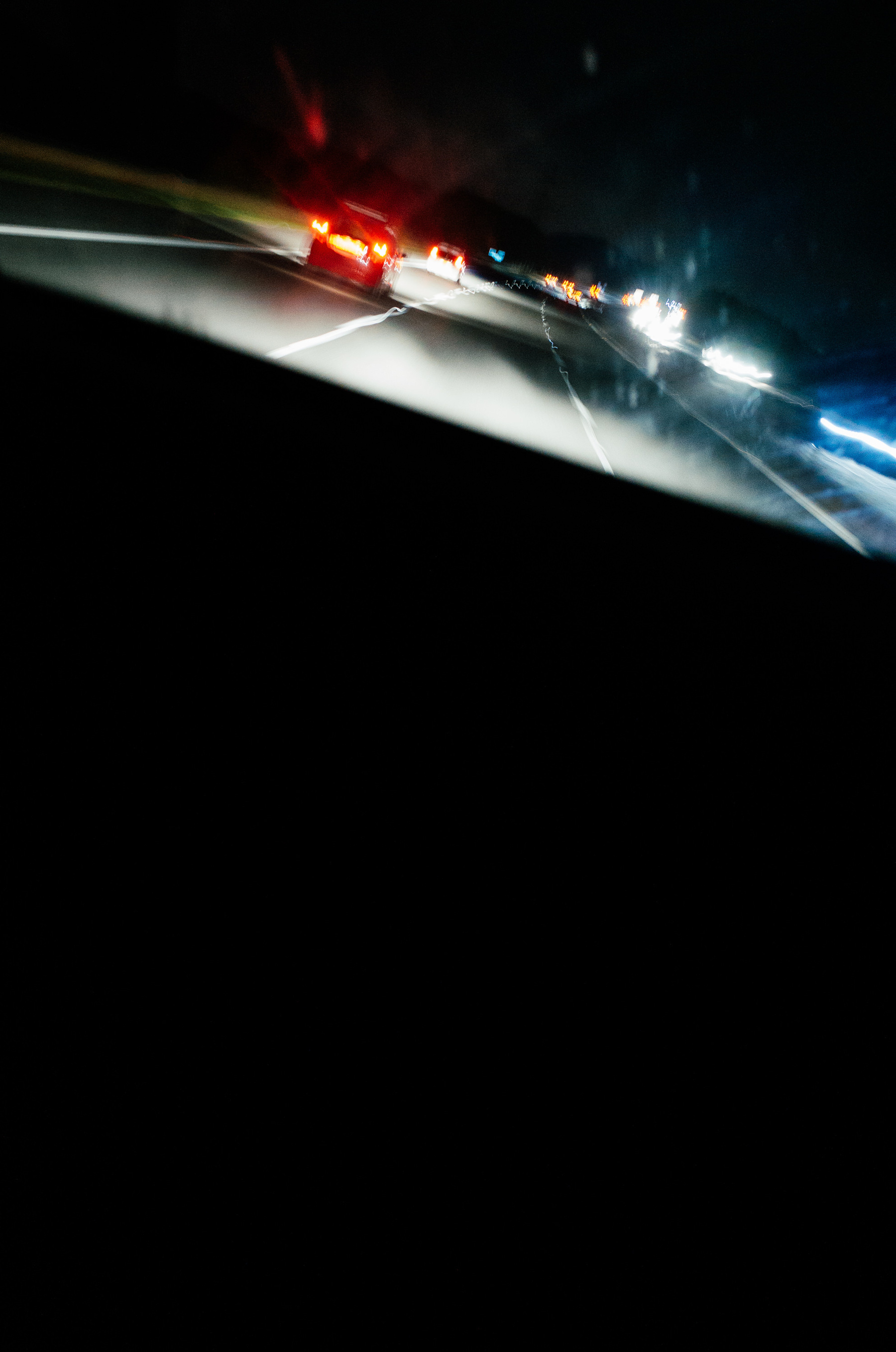 The top fifth of the frame shows cars and their brake lights driving away from the viewer on a motorway, the horizon tilted. You can see oncoming traffic in the top right hand corner. The rest of the image is completely black, obscured by the dashboard of the car that slices down and right from the top left corner of the frame