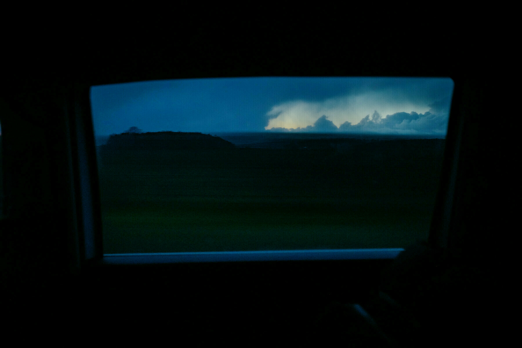 A car window frames a night time landscape, with the fading light of dusk seen through a hole in the storm clouds on the horizon. The interior of the car is dark and the landscape is blurred from by the speed of travel