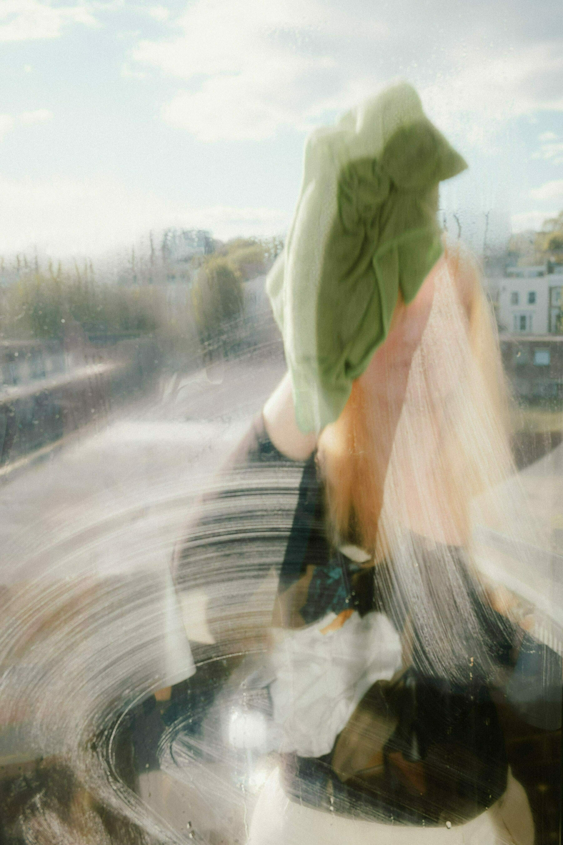 View through a window of a blonde woman washing the glass on the other side. The smeared soap suds and the green cloth in her hand are obscuring her face. She is standing on a balcony and residential buildings are visible in the background