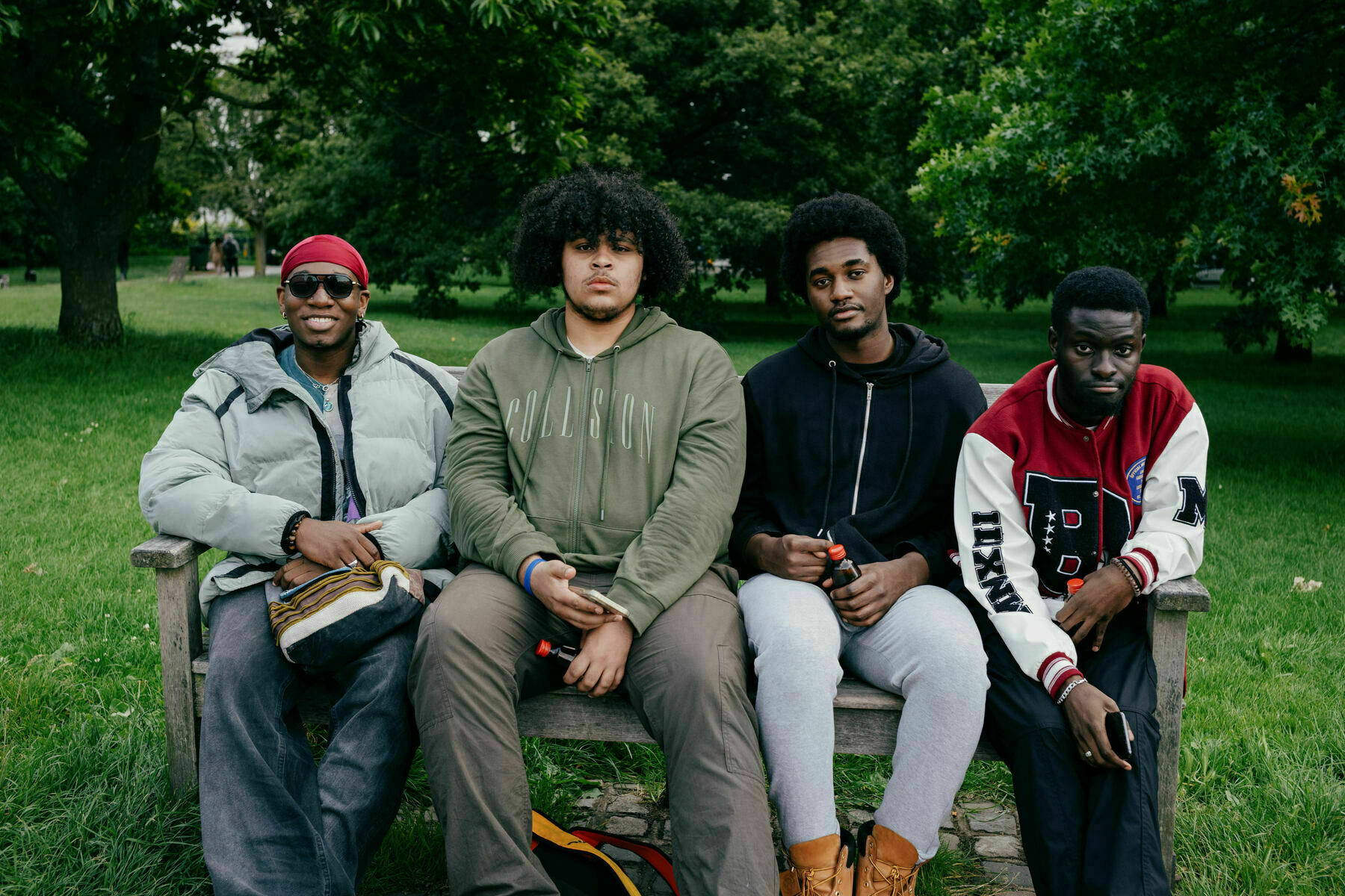 Portrait of four young men sitting on a park bench in Primrose Hill. From L to R they are wearing: grey puffer and red doo rag, khaki cargo pants and olive hoodie, light grey joggers and black hoodie, red, black and white varsity jacket and black cargo pants.