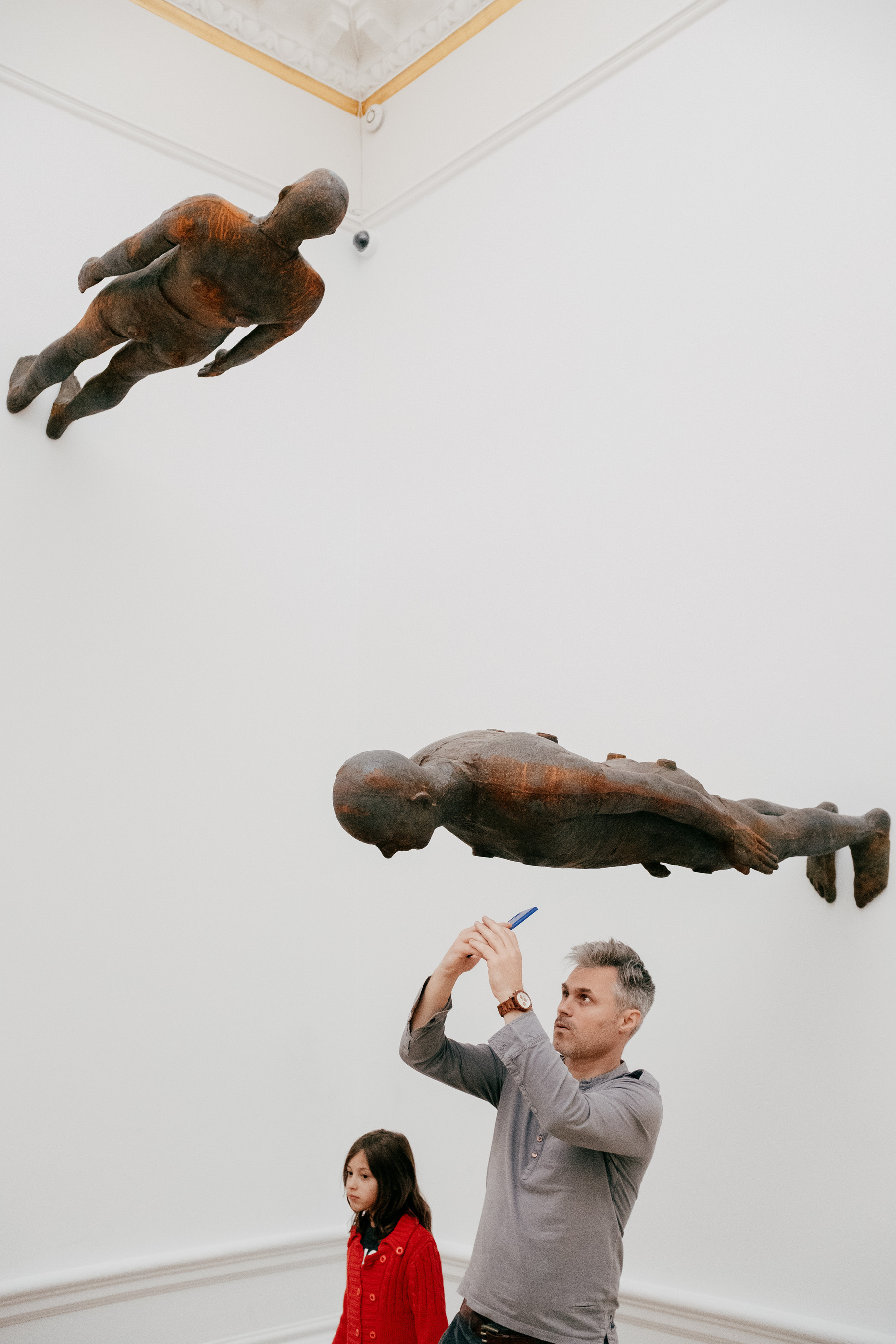 A man taking a photo of an Anthony Gormley sculpture of a man, that is affixed perpendicular to the gallery wall above his head. There is another sculpture of a man further up the adjacent wall and a young girl in a red dress is standing next to the man taking the photo.