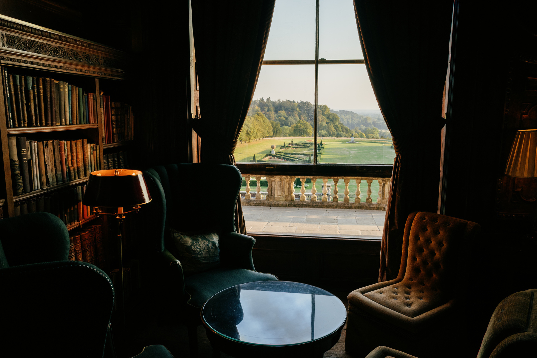 View out of the window of the library at Cliveden. There are armchairs on either side of the window in the foreground, and the grounds stretch towards the trees on the horizon, with a large ornamental garden visible to the left of the wide lawn