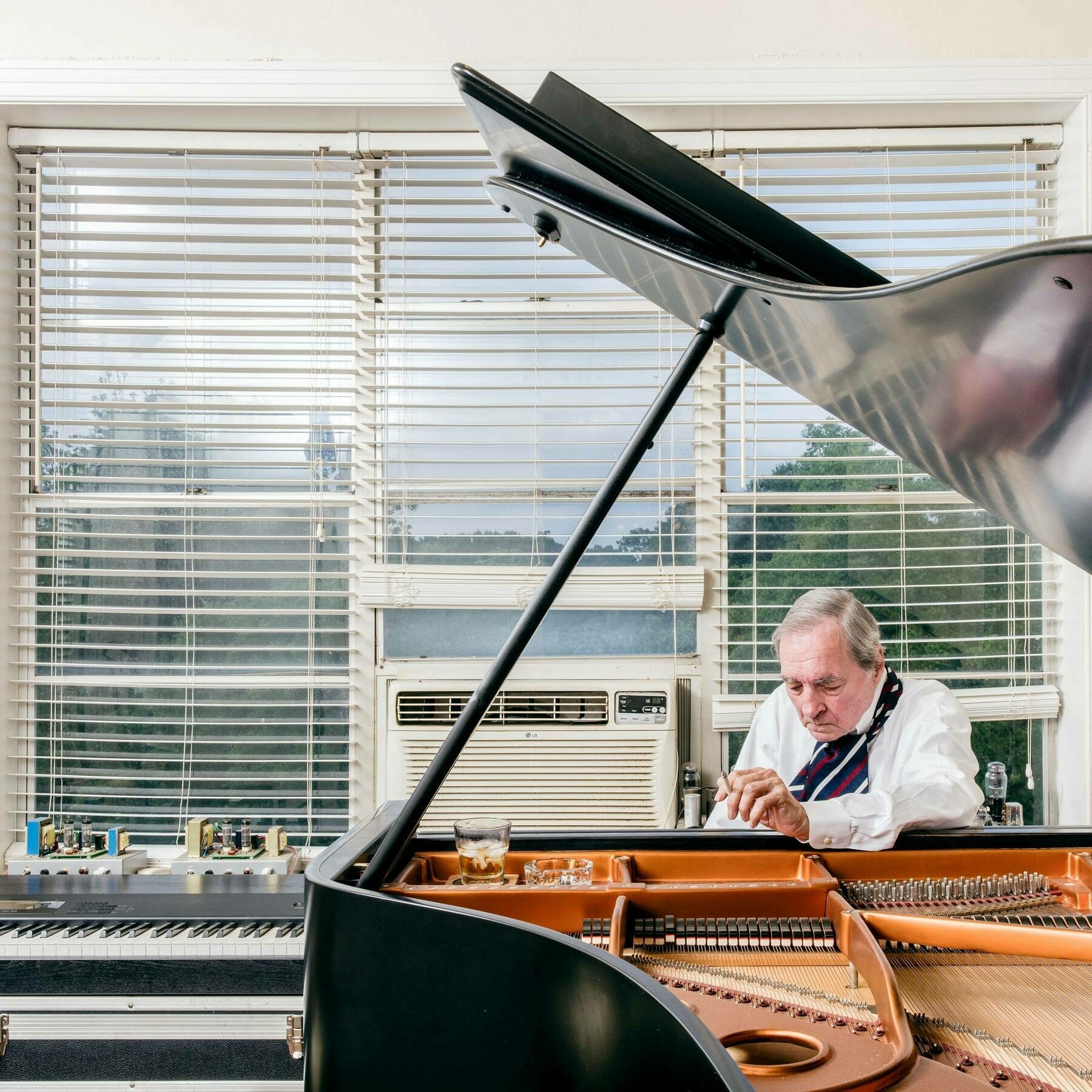 Portrait of photographer William Eggleston smoking at a piano with white metal blinds behind him.