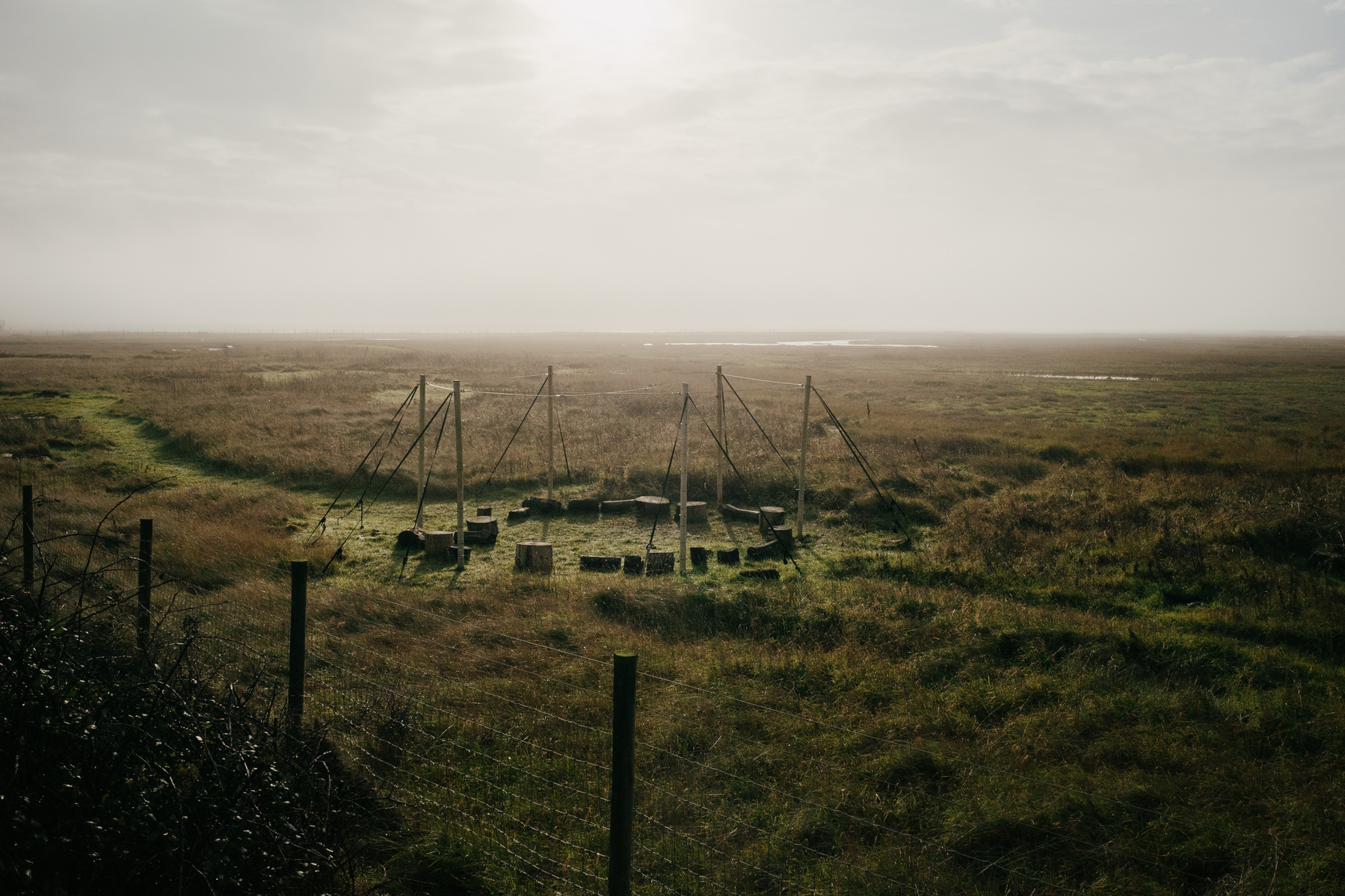 A misty landscape view of a minimal structure in open field at Rye Nature Reserve, with tall support poles and multiple tree trunk segments arranged in a circle as seating. The scene is bordered by wire fencing and stretches out to a hazy horizon where patches of water are visible in the distance.