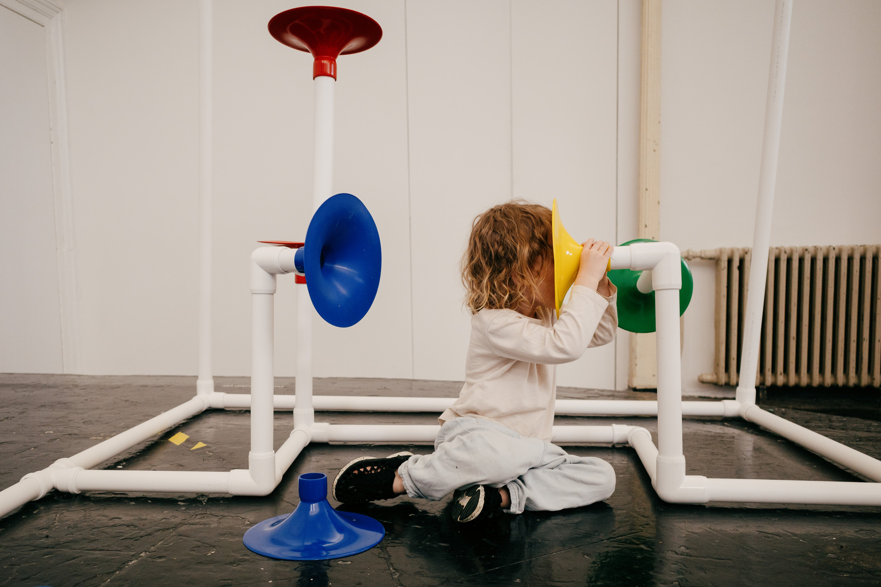 A small child hides their face in a trumpet-like 'speaker cone' that is part of a sculpture made of white plastic piping that surrounds them