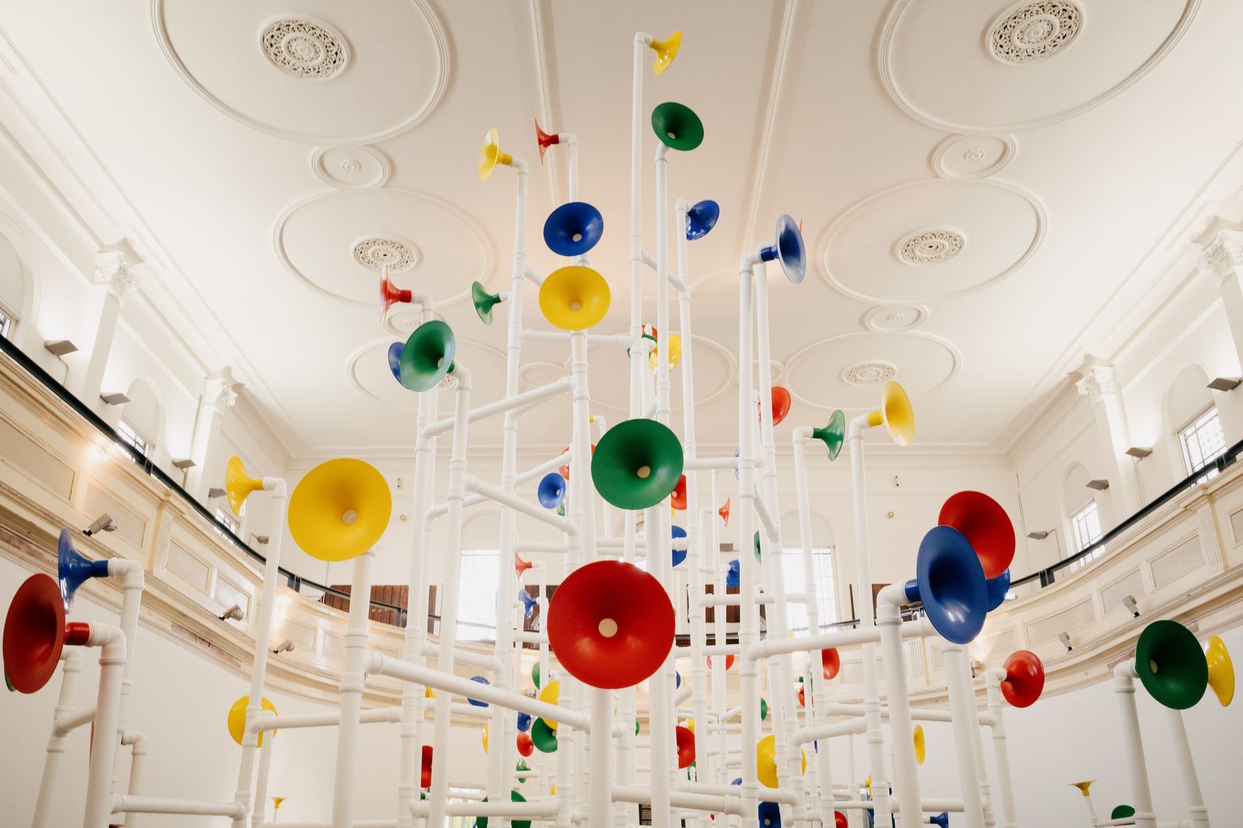 tower of white tubing with 'speakers' in different colours at the ends of various pipes. The structure is in a large, high-ceilinged, white room with multiple ceiling roses and large window high on the rear wall